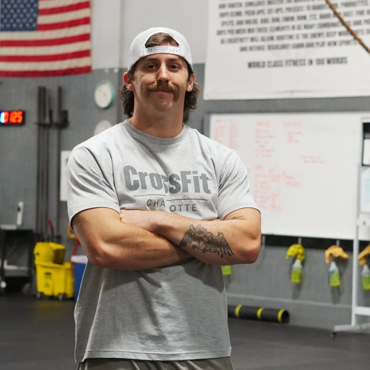 Bryson Delmonte standing with arms crossed in the gym, wearing a CrossFit Charlotte T-shirt and backwards cap.