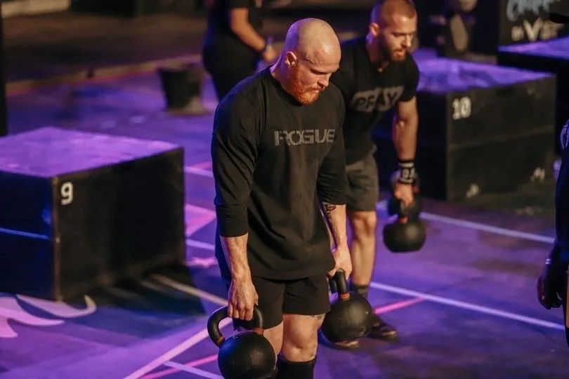 Taylor carrying two kettlebells at his sides on a competition floor lit in purple, another athlete behind him.