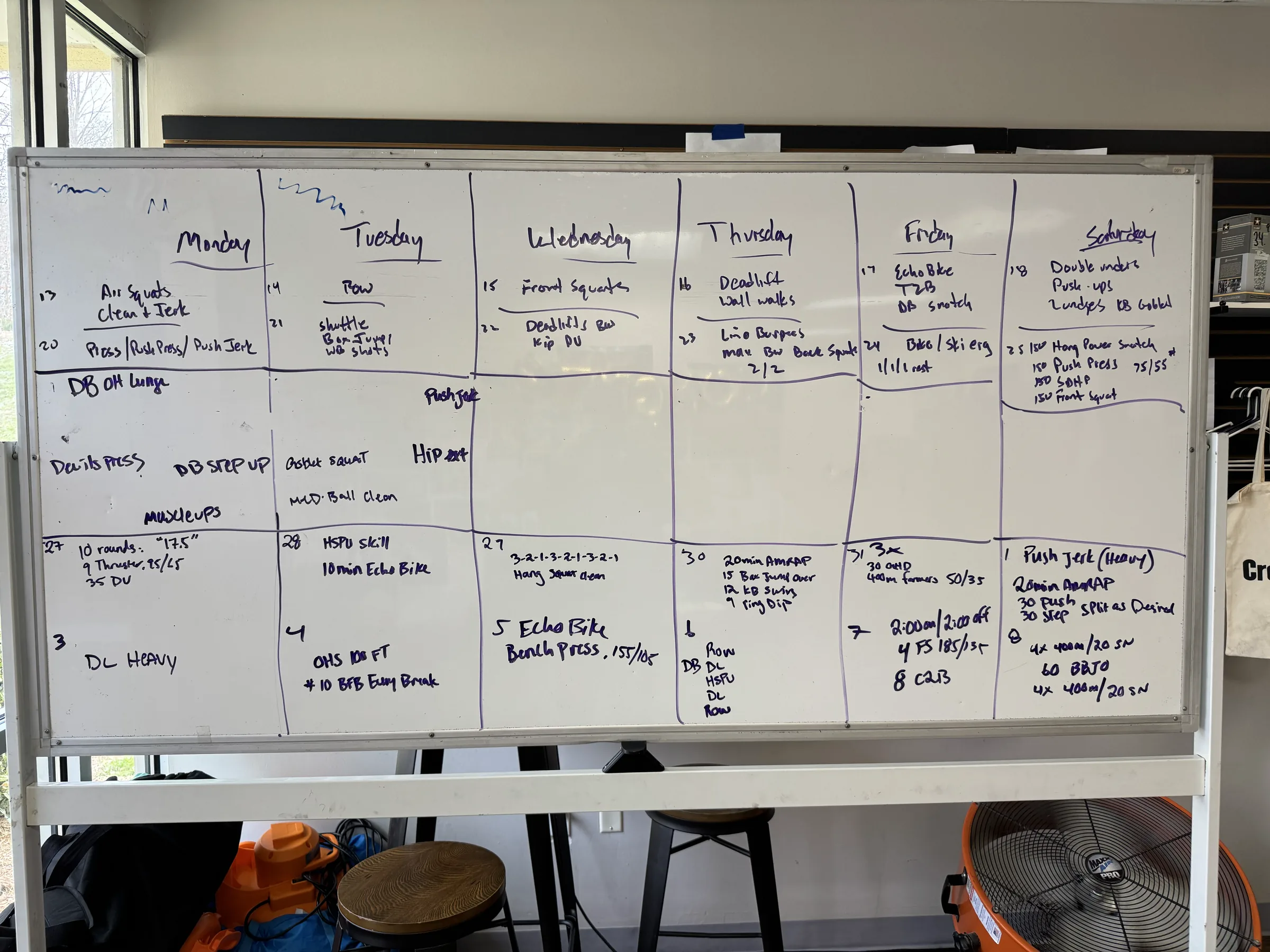 Whiteboard grid of a week of training, Monday through Saturday, filled in with handwritten lifts and metcons.