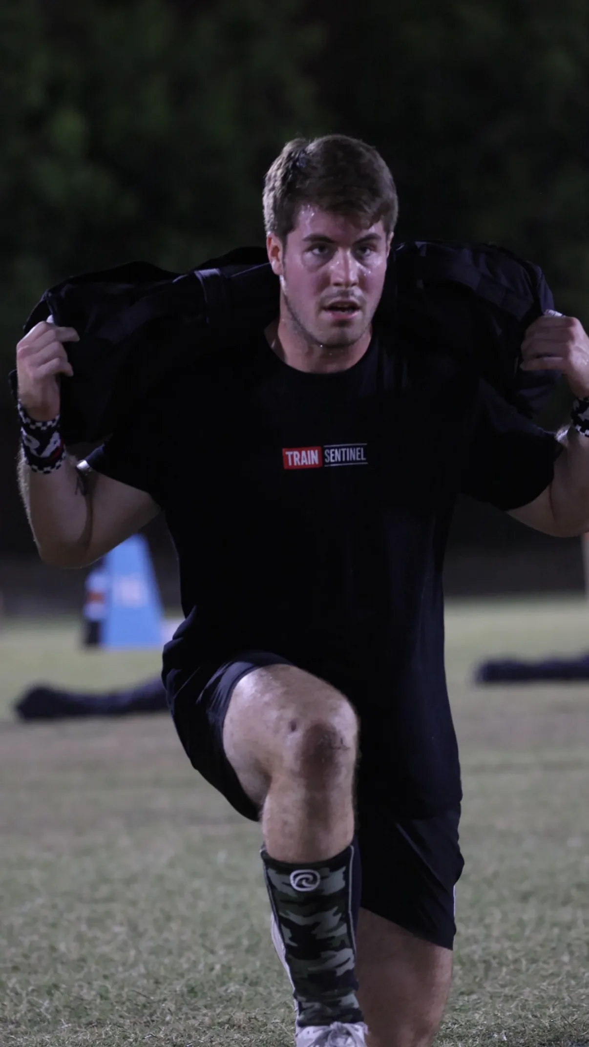 Will Plummer lunging with a sandbag across his shoulders on an outdoor field.