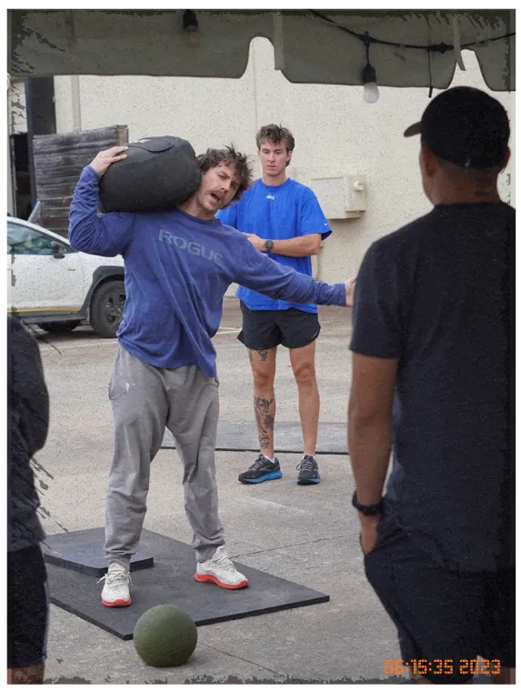 Athlete carrying a sandbag on one shoulder across the gym floor while coaches look on.