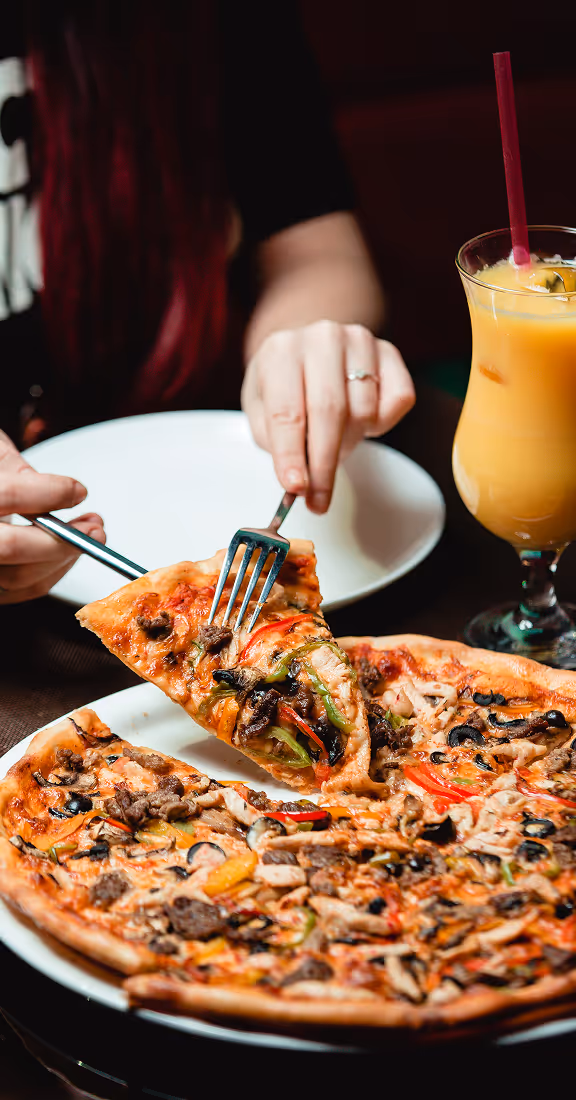 Person holding a slice of vegetable and meat pizza with a fork and knife, with a glass of orange juice nearby.