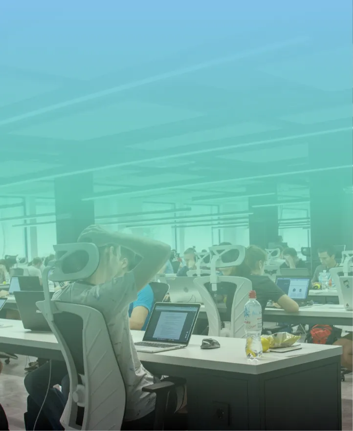 Open office space with multiple people sitting at desks working on laptops, featuring white ergonomic chairs and large windows in the background.