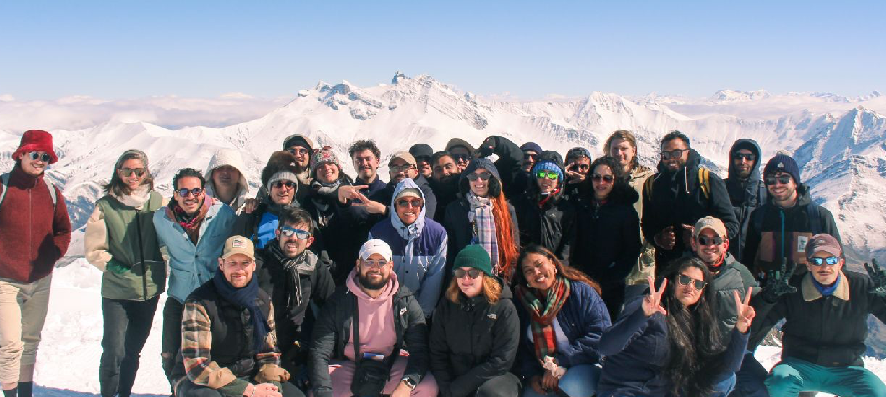 Un grand groupe diversifié de personnes vêtues de vêtements d'hiver, posant avec le sourire sur une montagne enneigée, avec des sommets couverts de neige et un ciel bleu clair en arrière-plan.