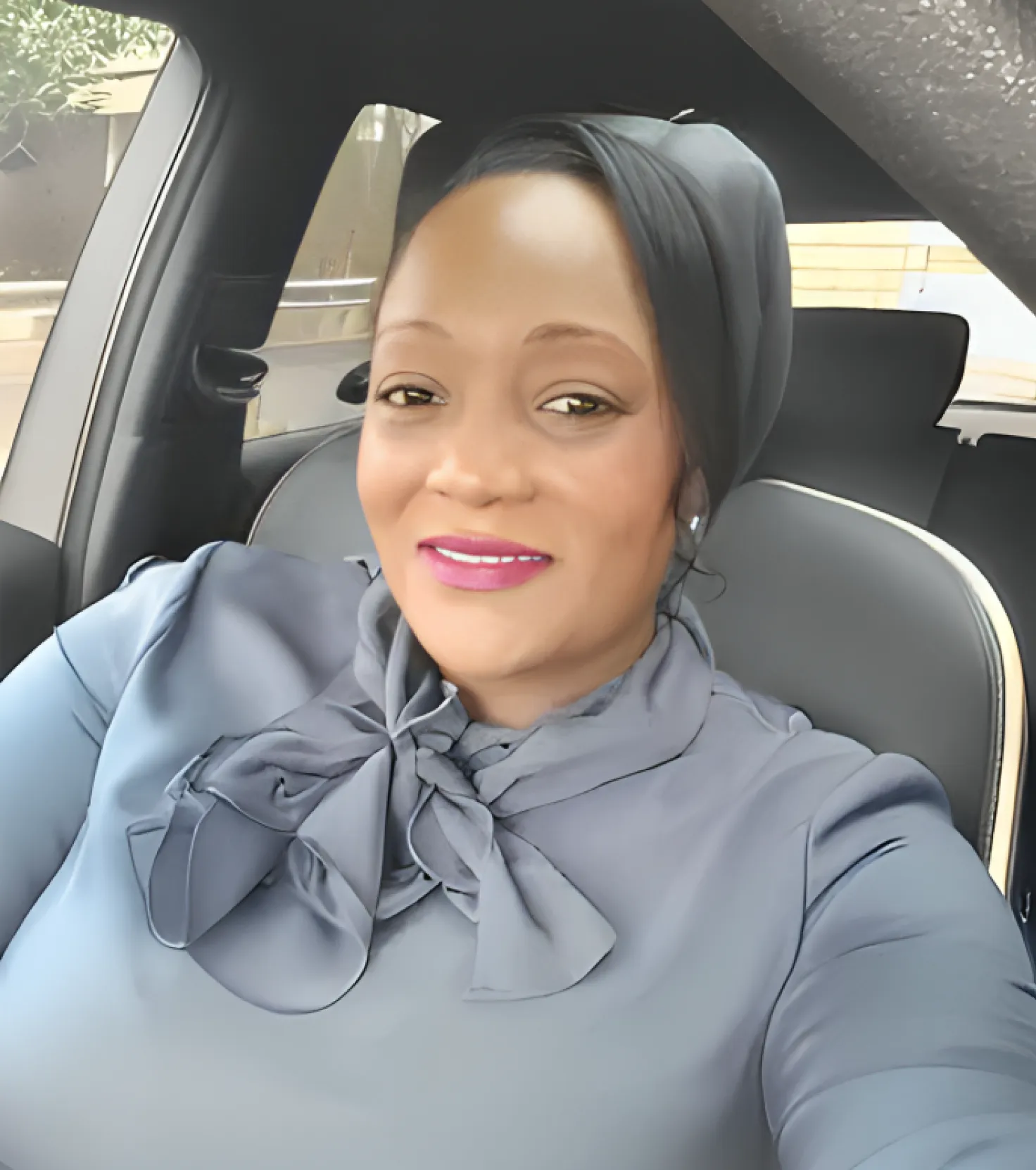 Smiling woman wearing a black headscarf and a gray blouse with a bow tie seated inside a car.