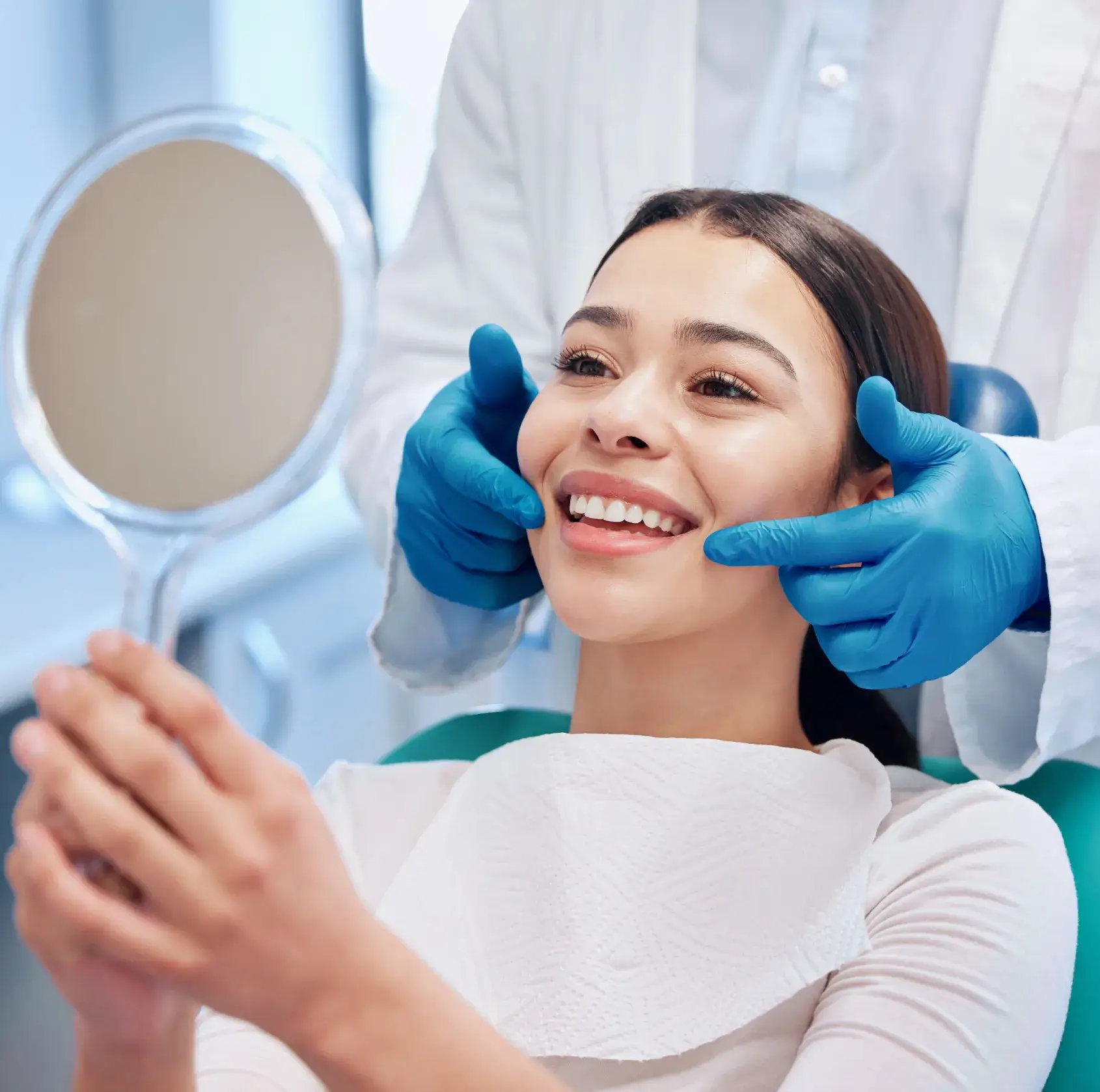 A person in a dental chair smiles while holding a mirror, with a dentist's hands adjusting their cheeks.