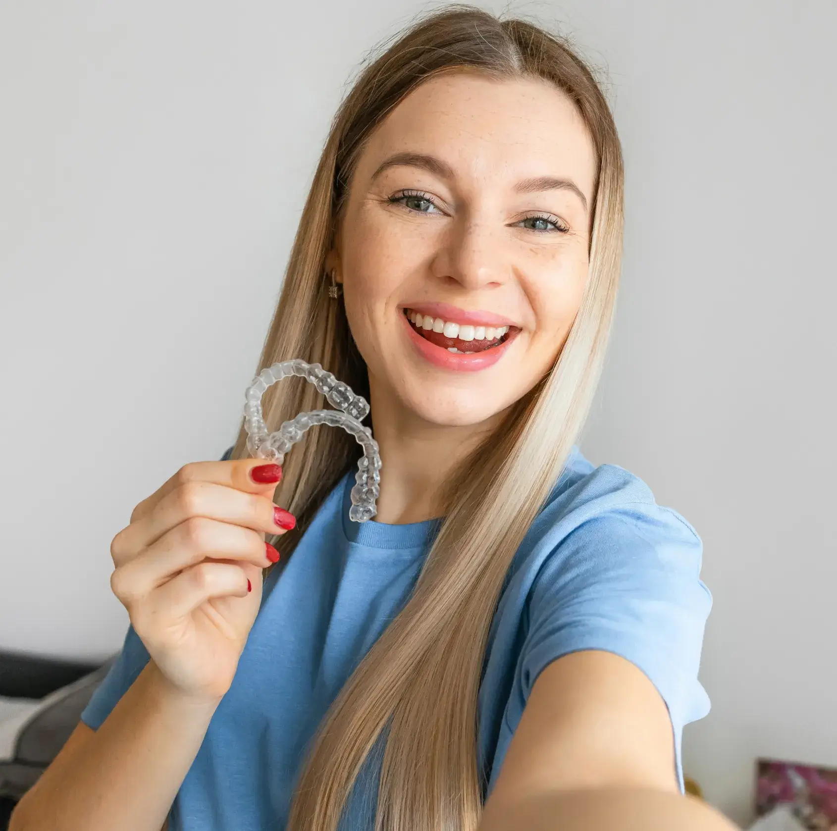 Smiling woman with long blonde hair wearing a blue shirt holds up clear dental aligners.