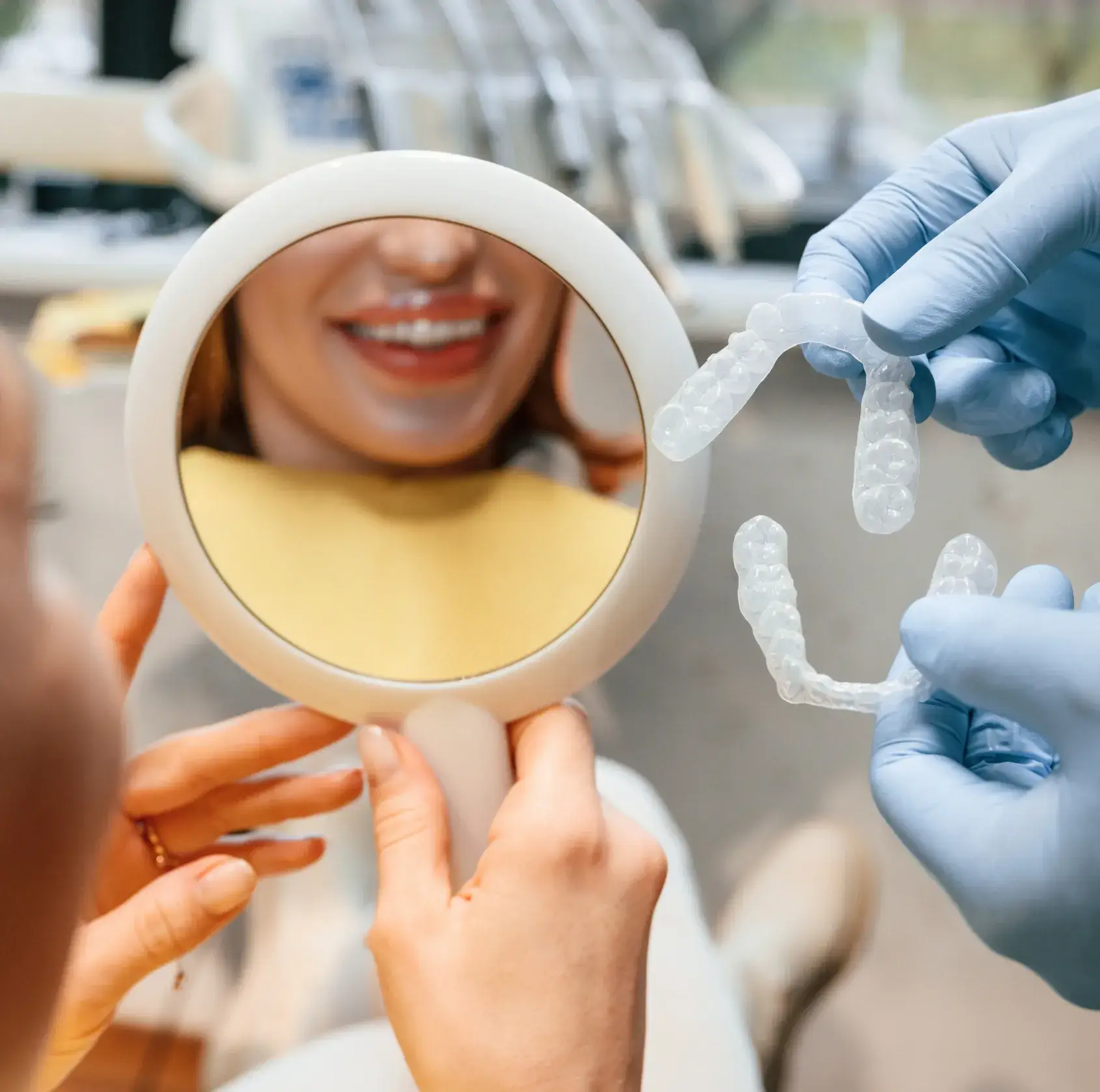 A person holds a mirror, smiling, while a dentist shows clear dental aligners with gloved hands.