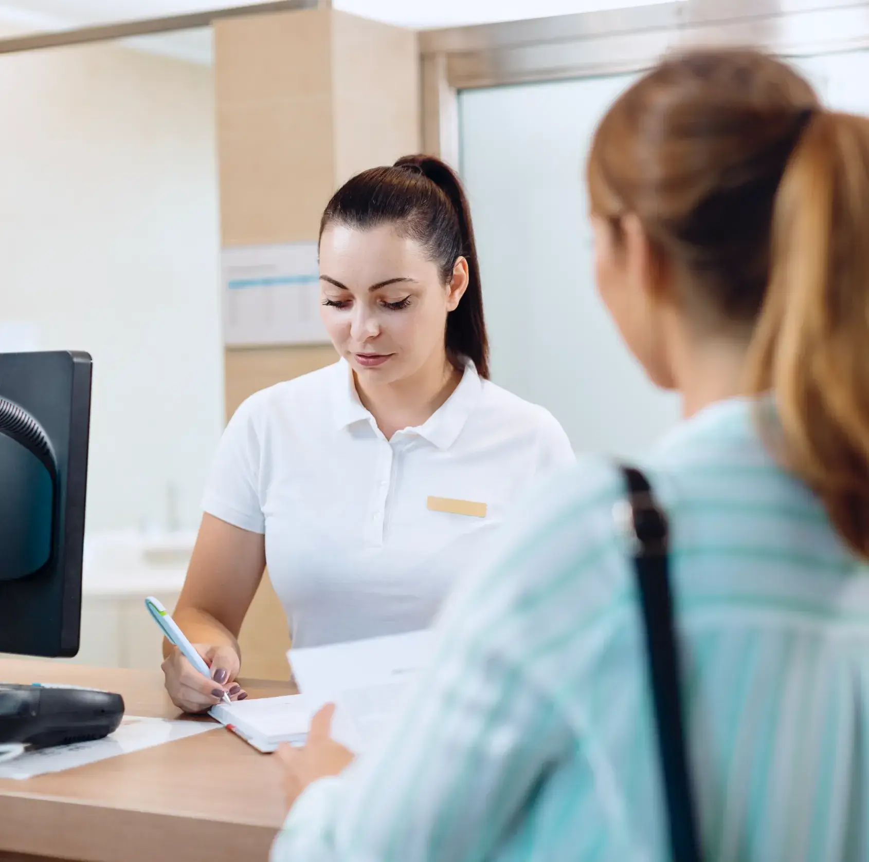 Receptionist in white uniform writing on a clipboard at a desk while a woman in a striped shirt waits.