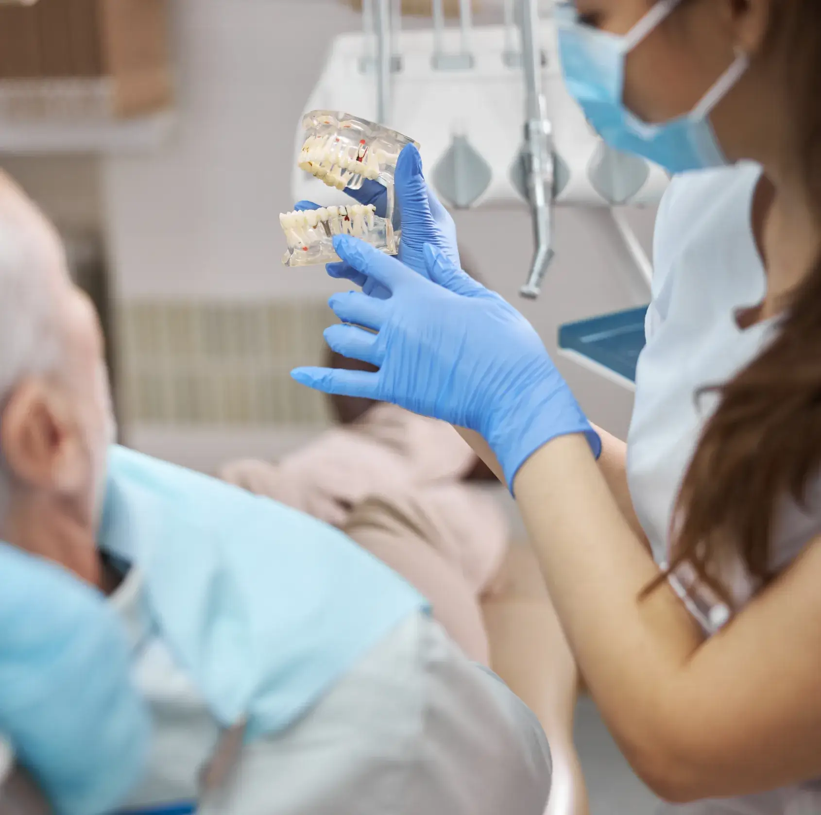 Dentist holds a dental model while talking to a seated patient in the examination room.
