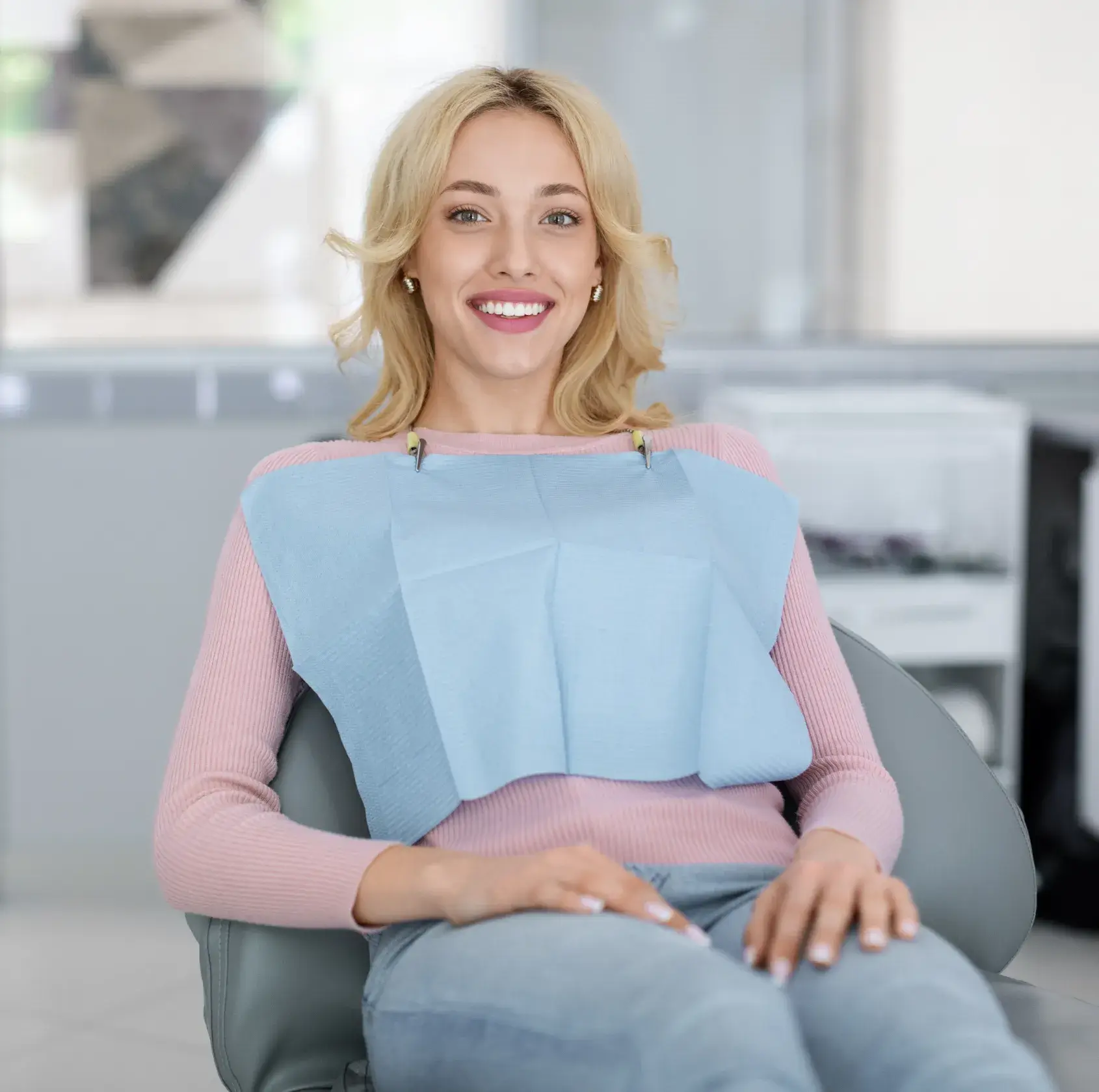 Smiling young woman wearing a pink sweater and blue dental bib sitting in a dental chair.