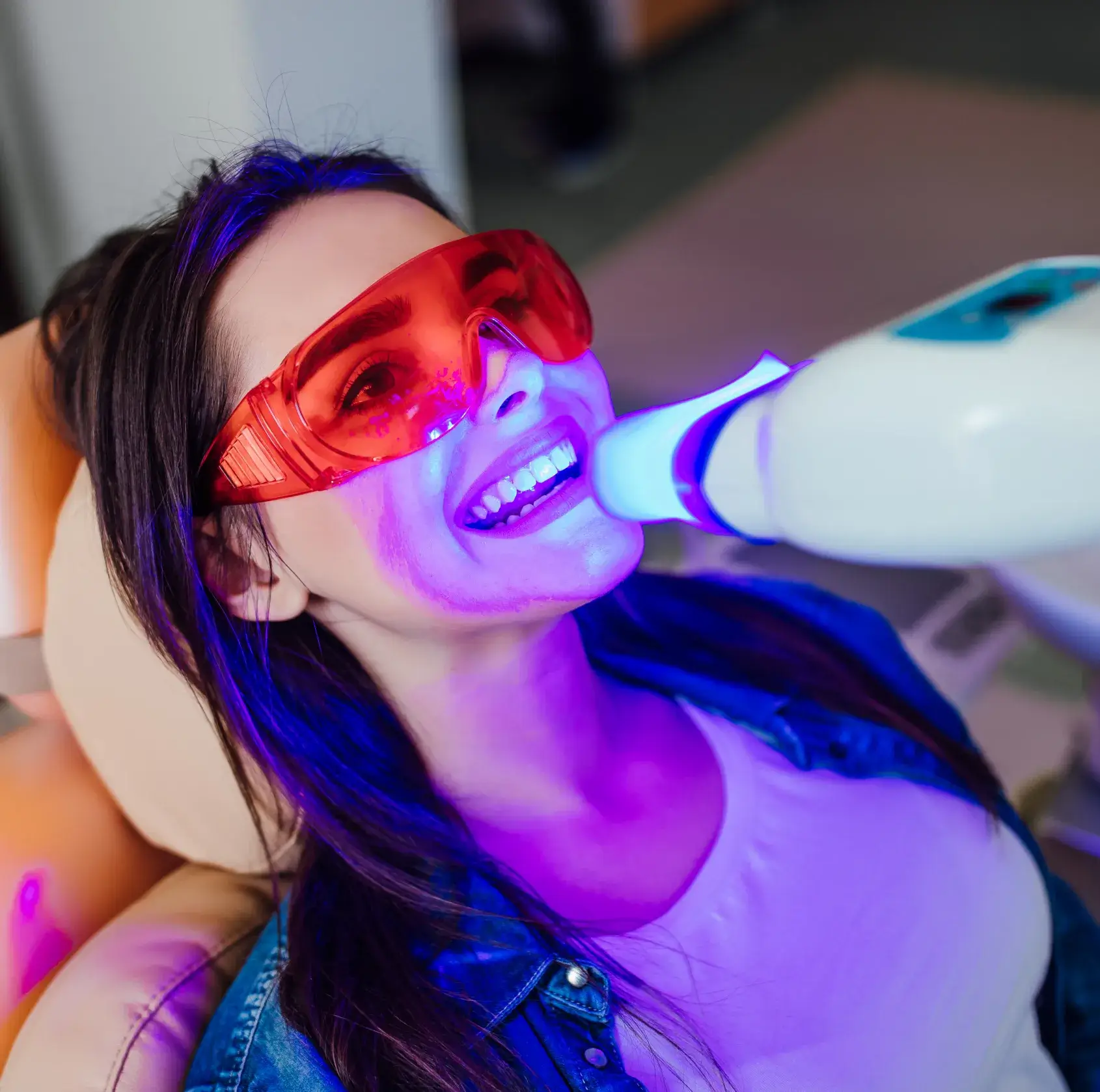 Woman wearing red protective glasses undergoing a blue light teeth whitening treatment.