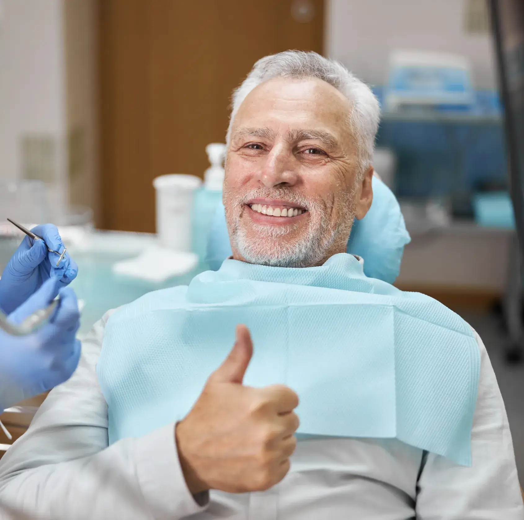 Smiling elderly man in dental chair giving a thumbs up, with dentist's gloved hands holding dental tools nearby.