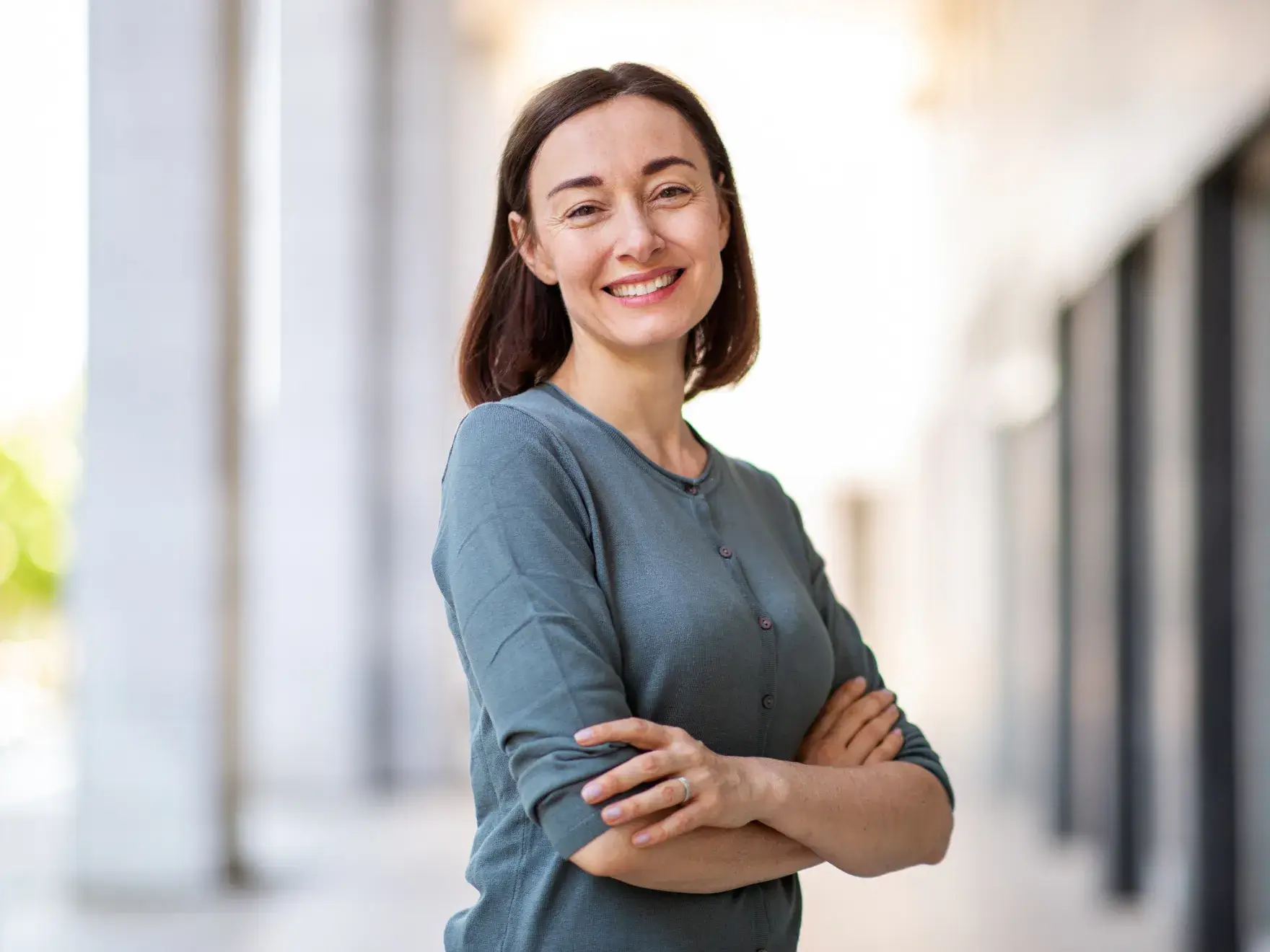 A woman smiles while standing outdoors with her arms crossed, wearing a long-sleeved gray shirt.