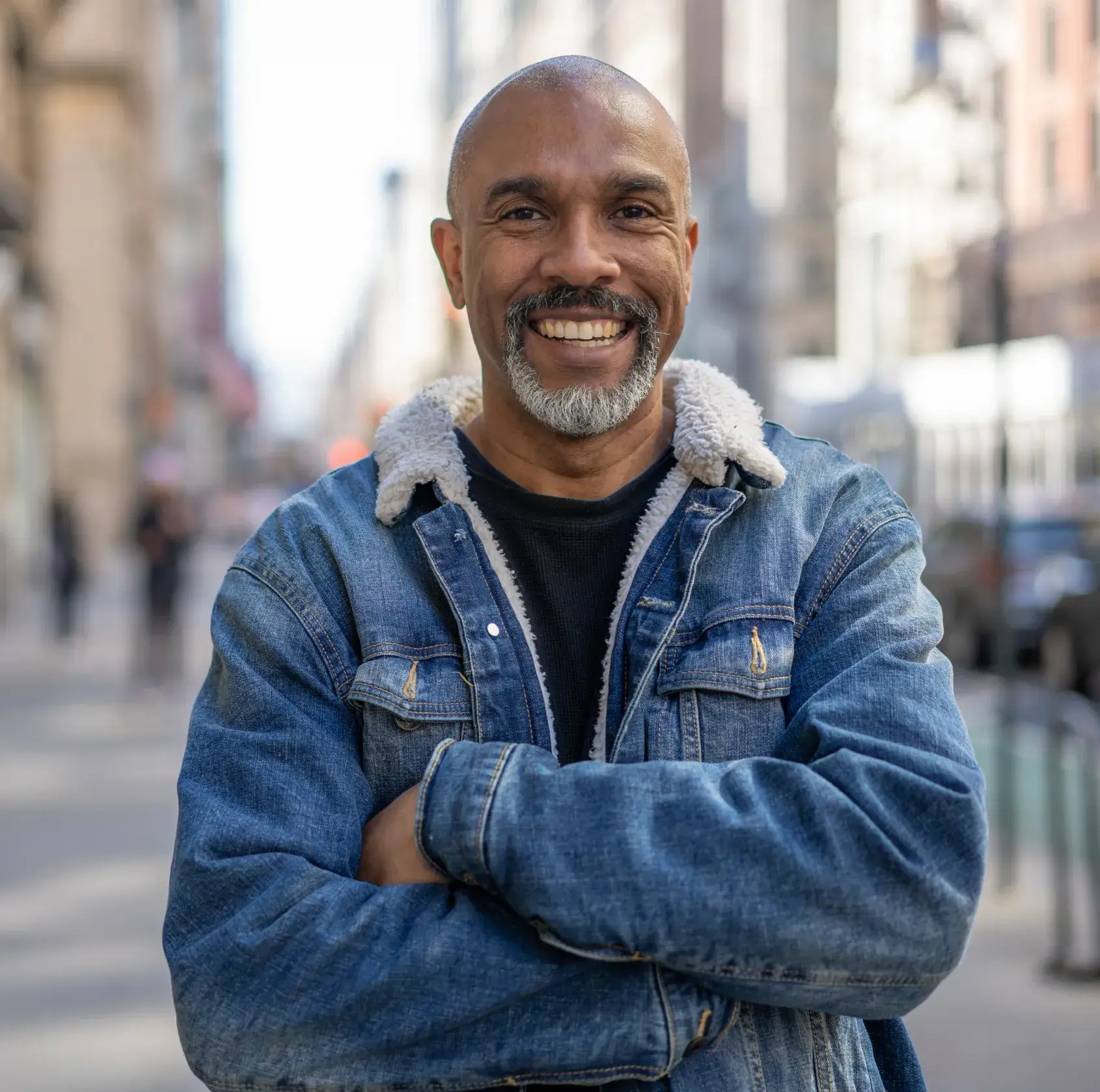 A man in a denim jacket smiles confidently with his arms crossed on a city street.