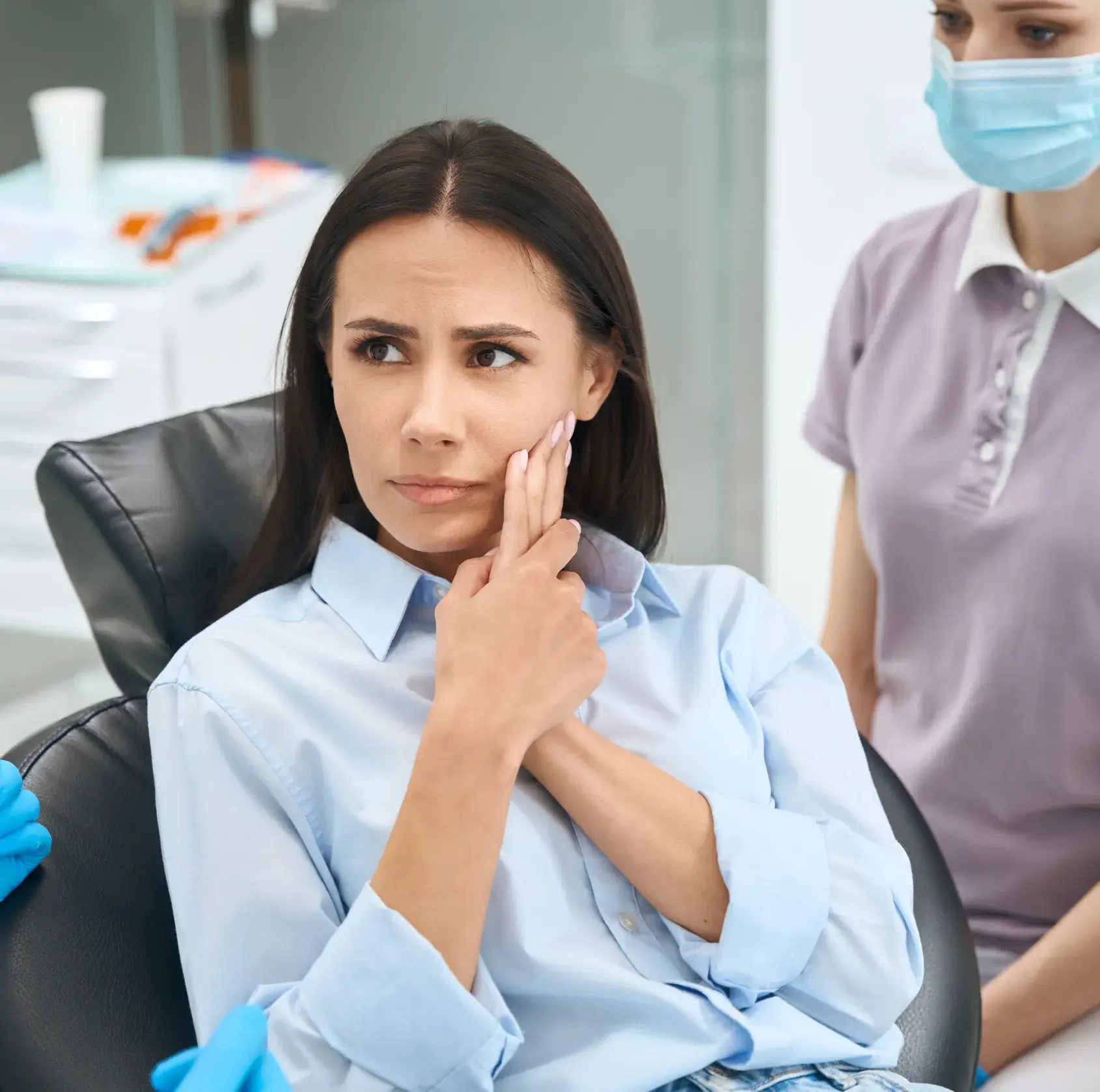Woman sitting in a dental chair holding her cheek in pain, with a masked dental professional nearby.