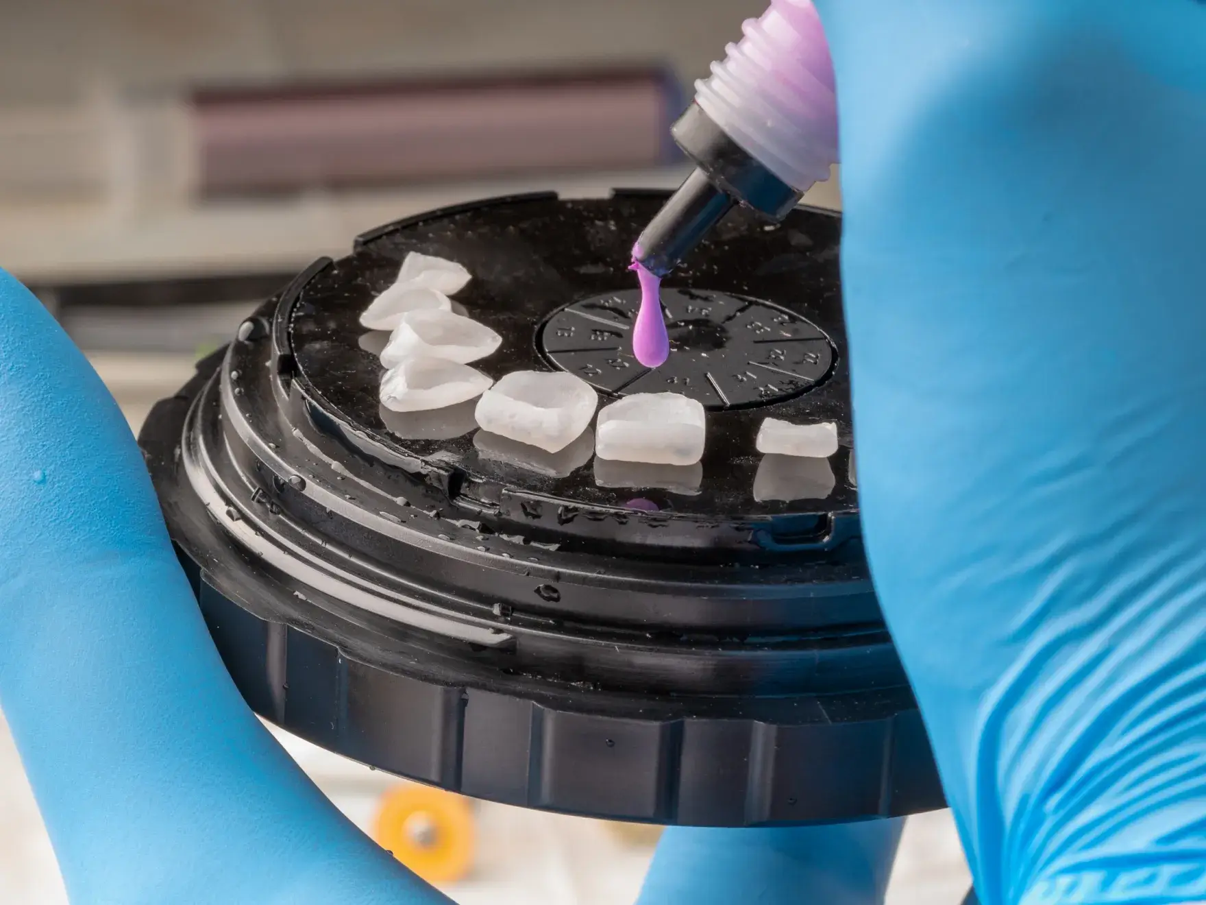 A person in blue gloves applies pink liquid to dental veneers on a circular tray.