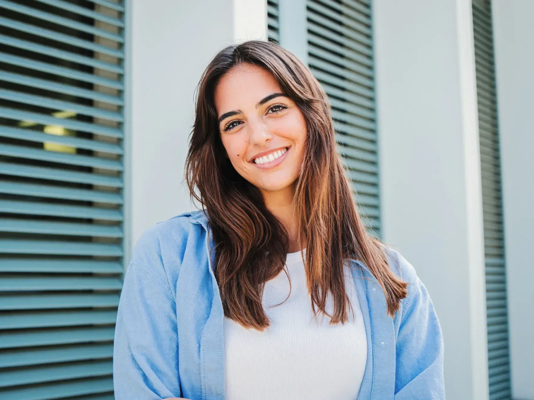 Smiling woman with long brown hair wearing a white top and blue jacket standing in front of a building with green shutters.