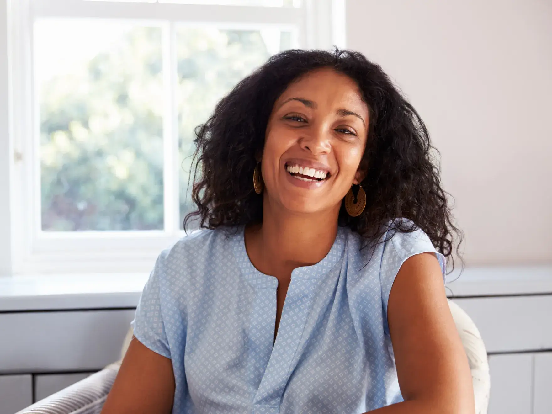 A woman with curly hair smiles while sitting in front of a window.