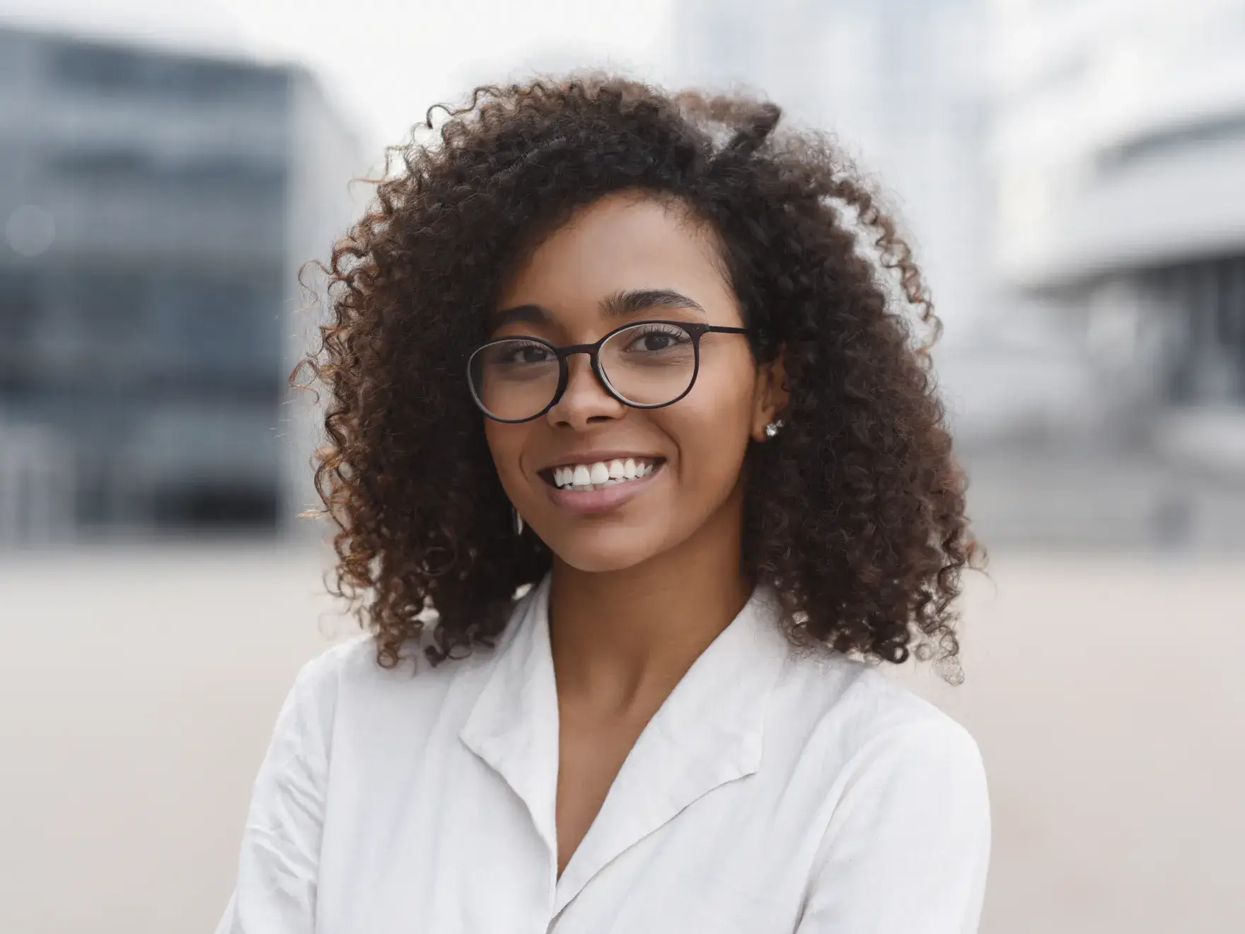 A person with curly hair and glasses smiles while standing outside.