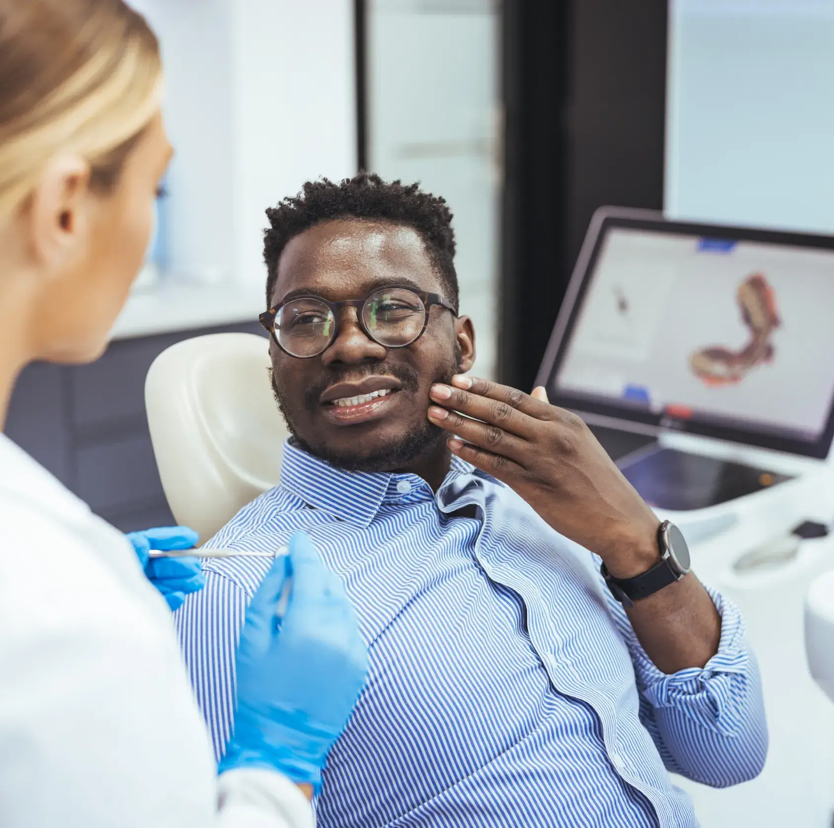 A dentist talks to a patient sitting in a dental chair, who is touching his face.