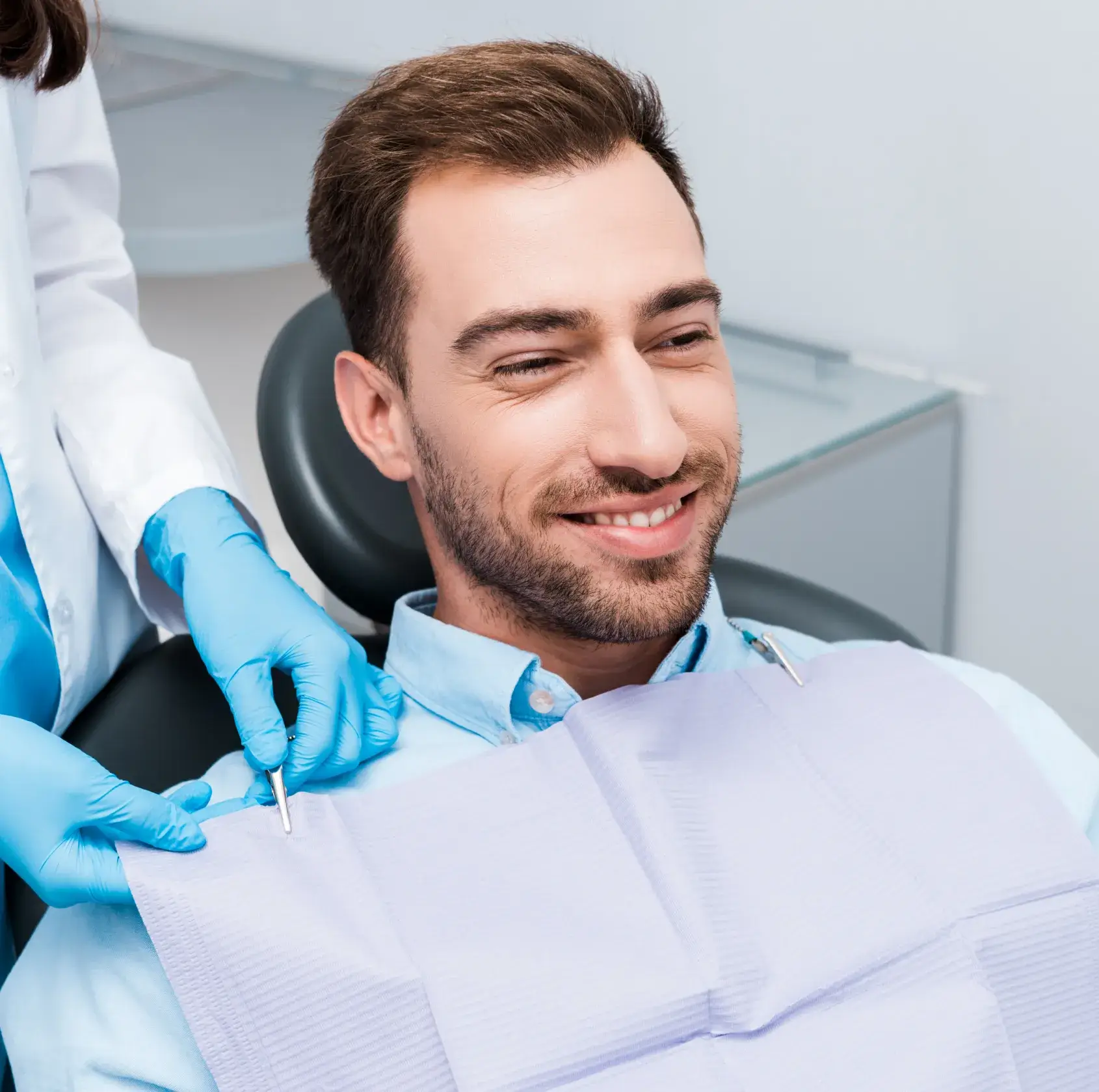 A dentist prepares a patient for treatment using dental tools while he sits and smiles.
