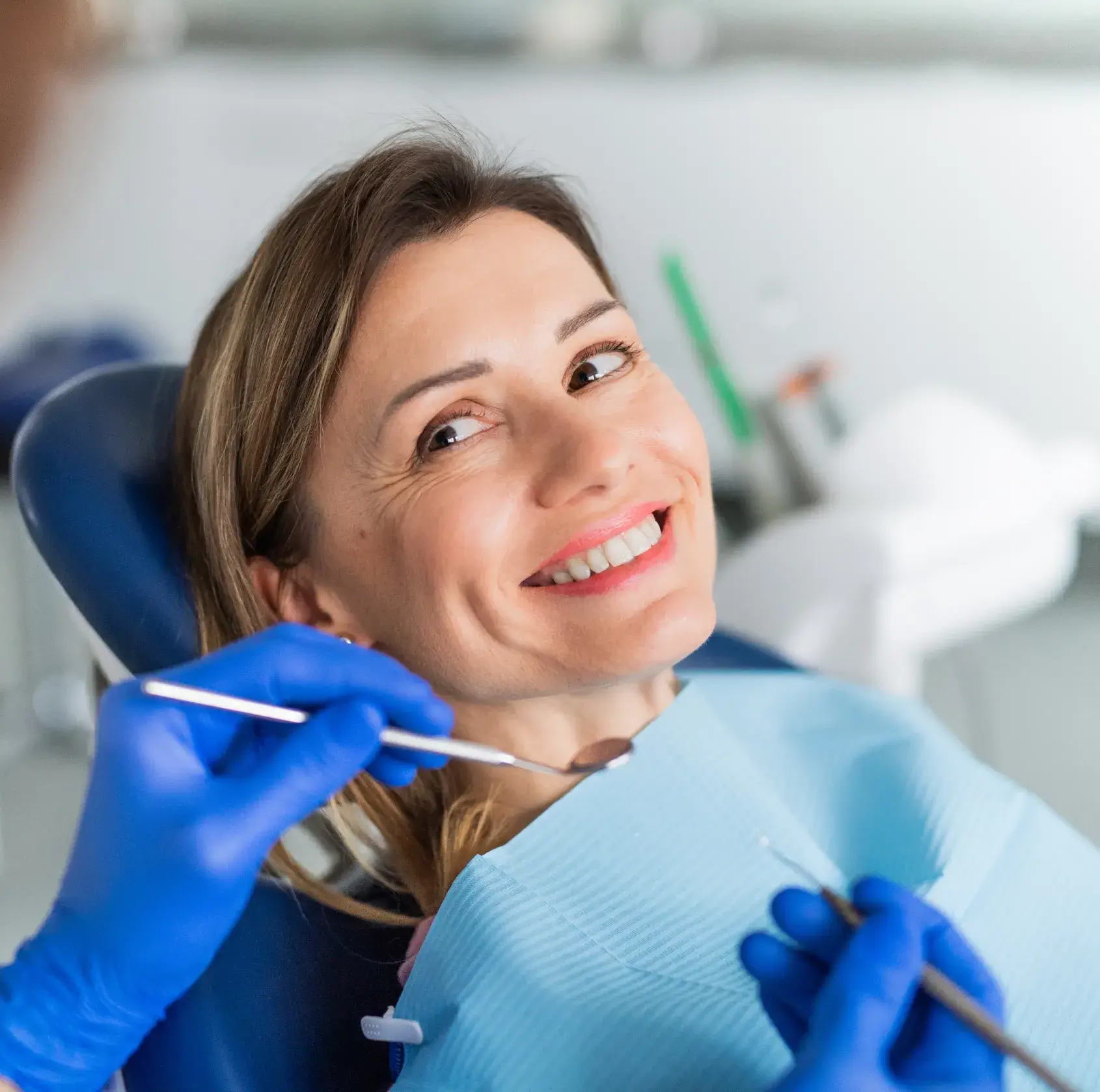 A woman smiles in a dental chair while a dentist examines her teeth with tools.