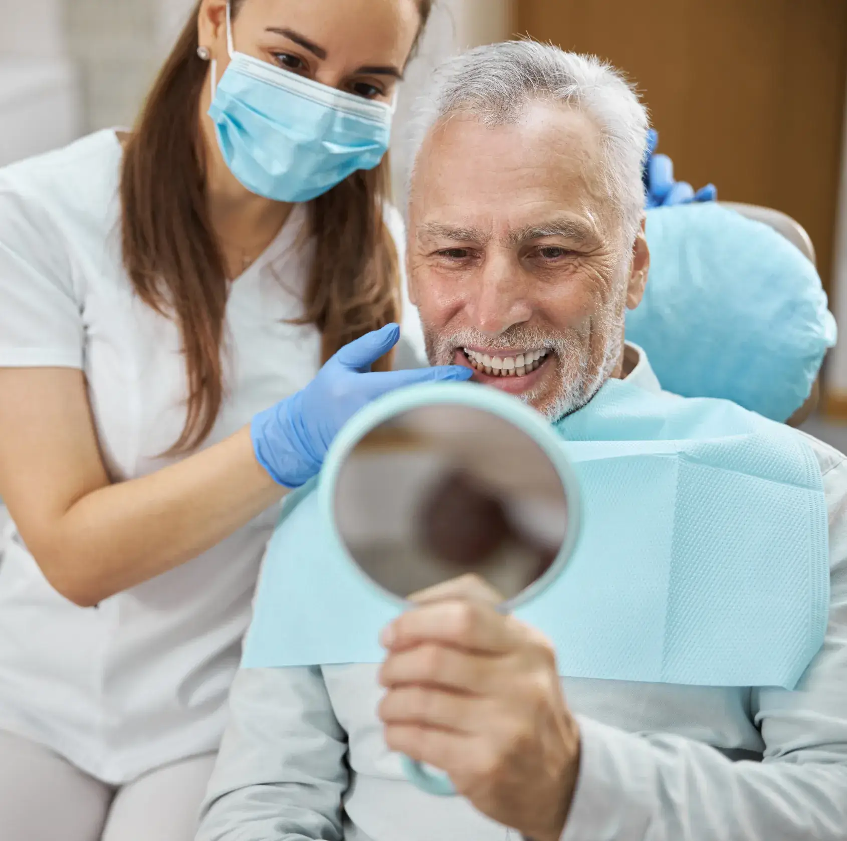 Elderly man in dental chair smiling while looking at his teeth in a handheld mirror, with a masked female dentist wearing blue gloves pointing at his teeth.
