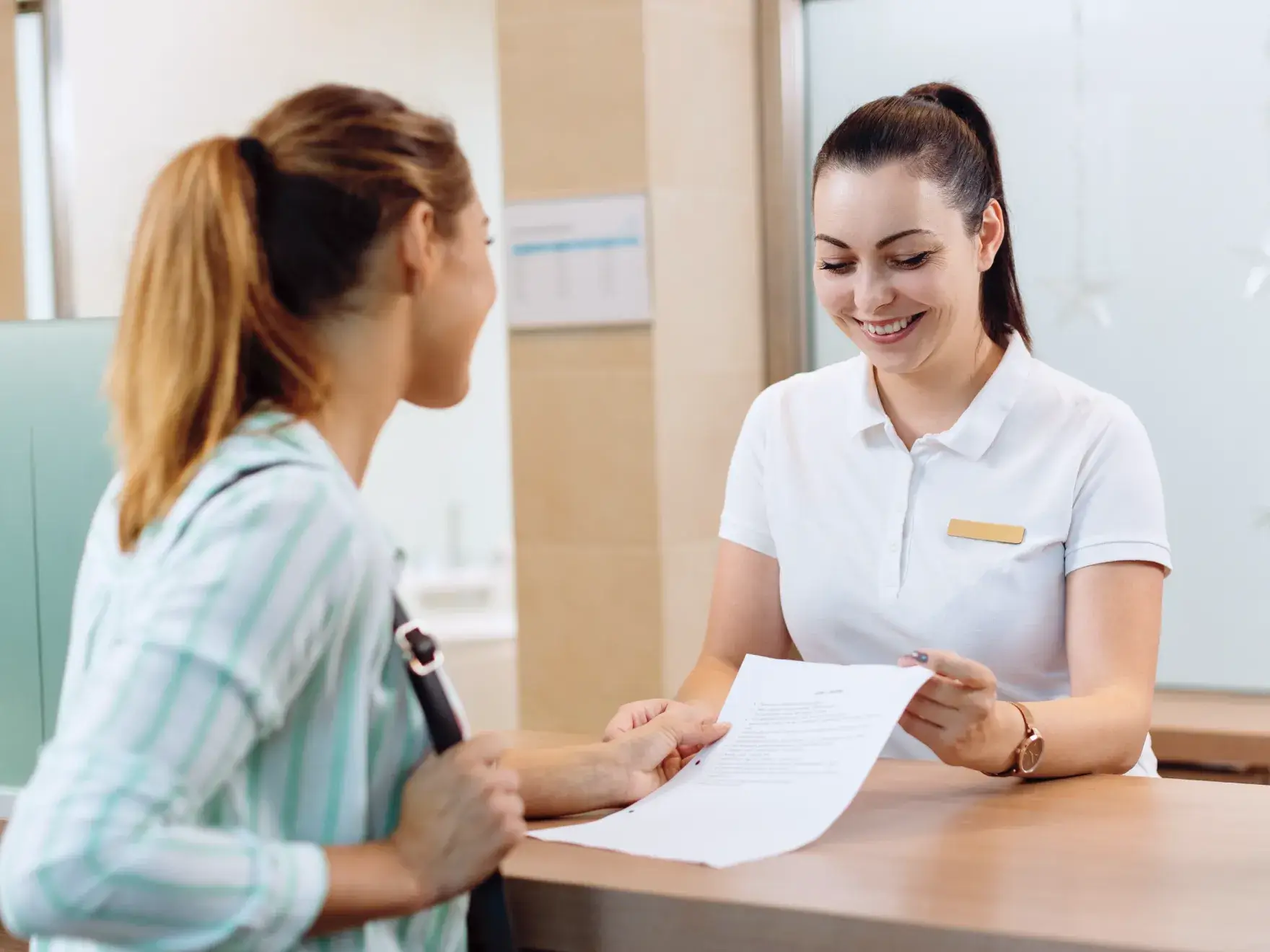 Receptionist smiling and handing a document to a woman across the desk in an office setting.
