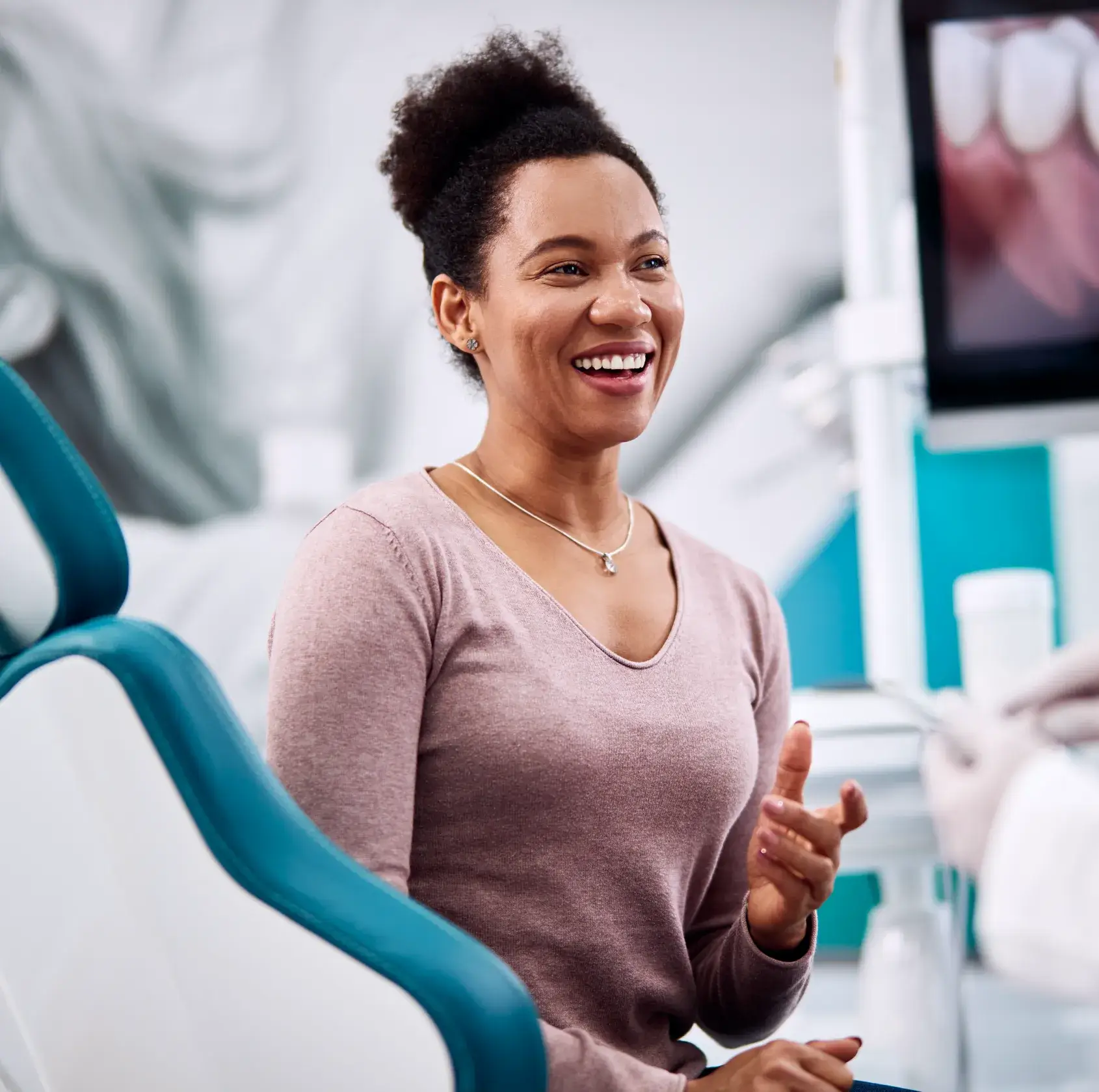 A woman sits in a dental office chair, smiling and looking towards someone.
