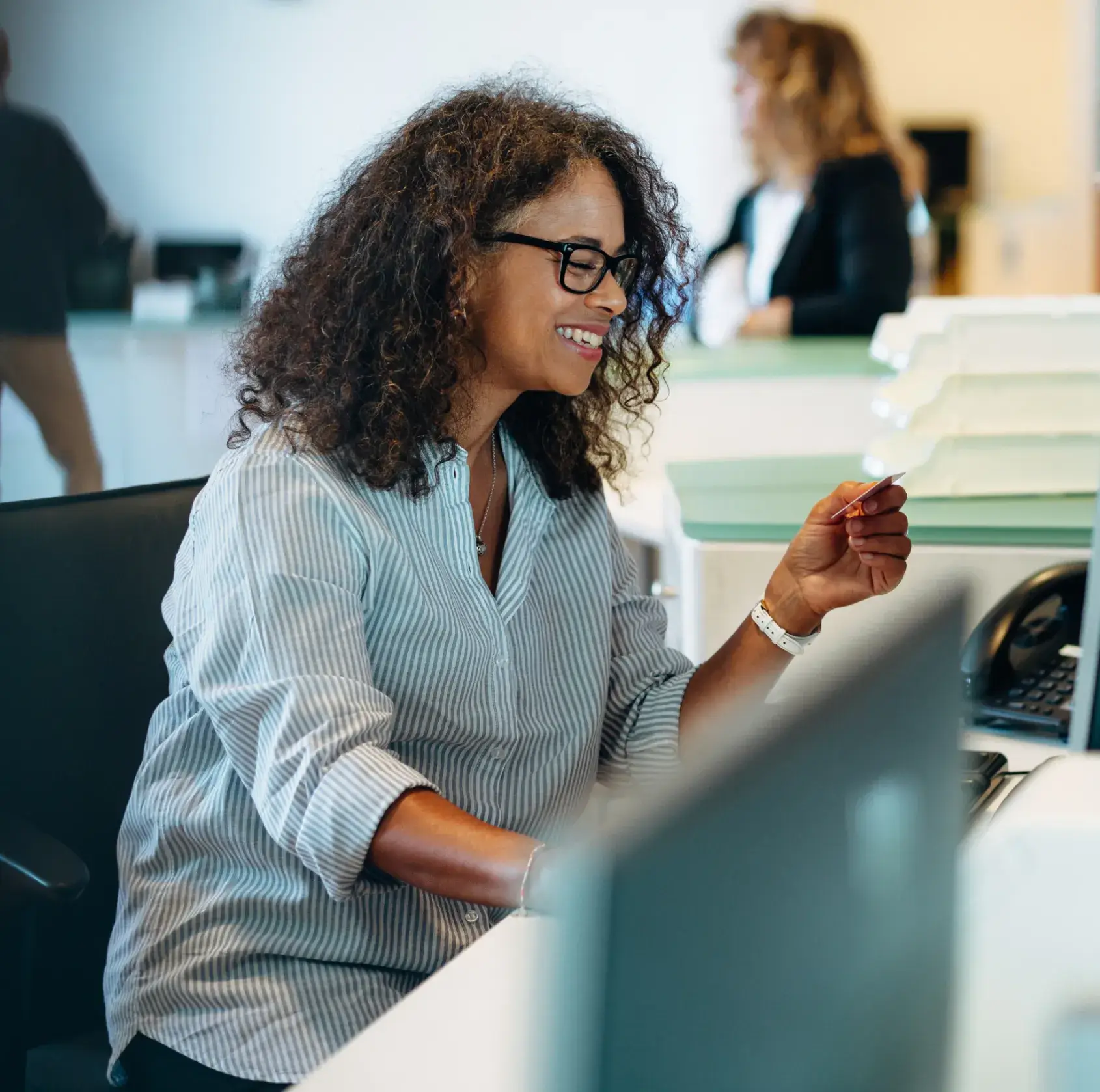A woman wearing glasses sits at a desk, smiling, while holding a card in her hand.