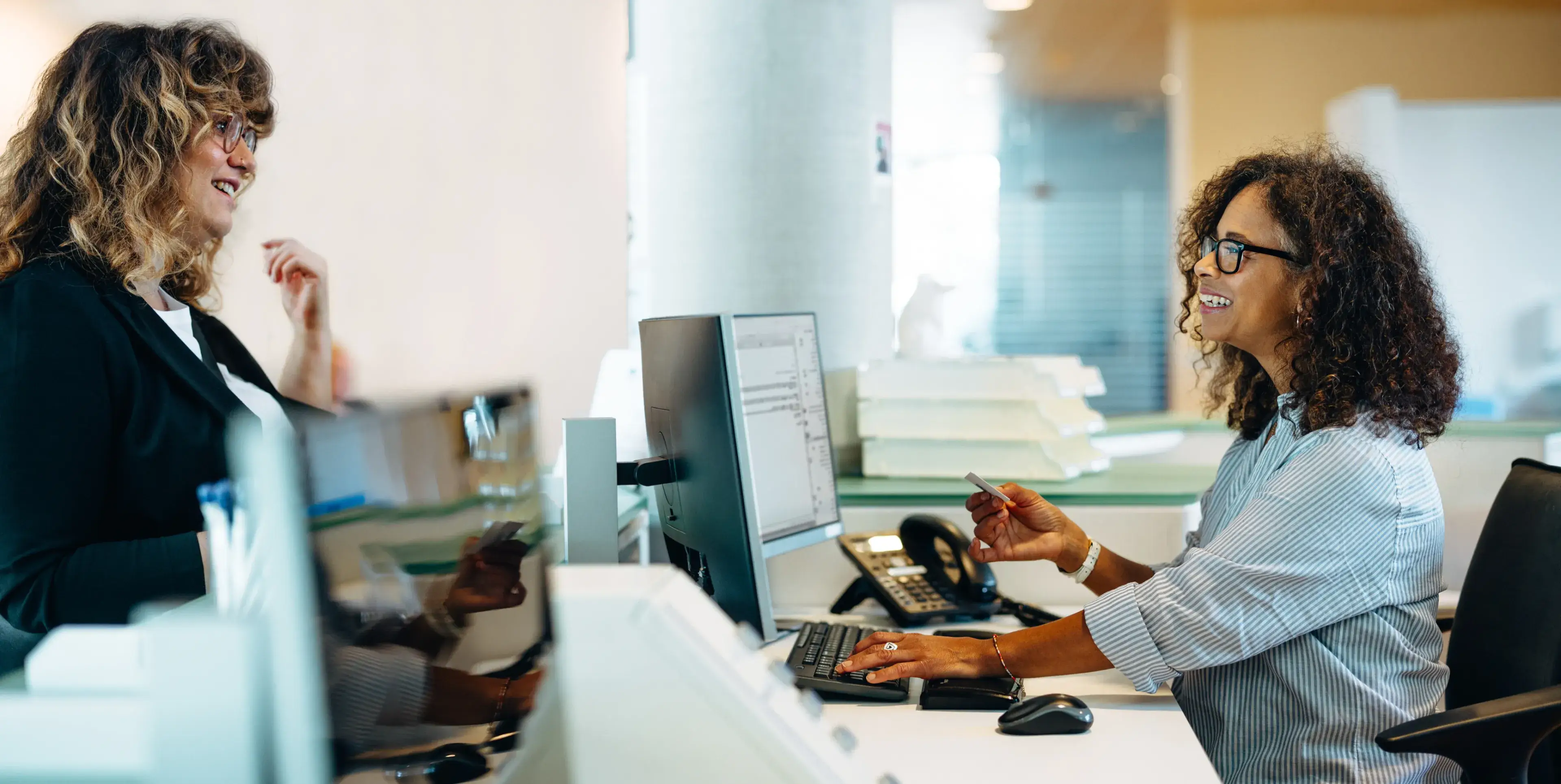Two people talking at an office desk with computers and phones.