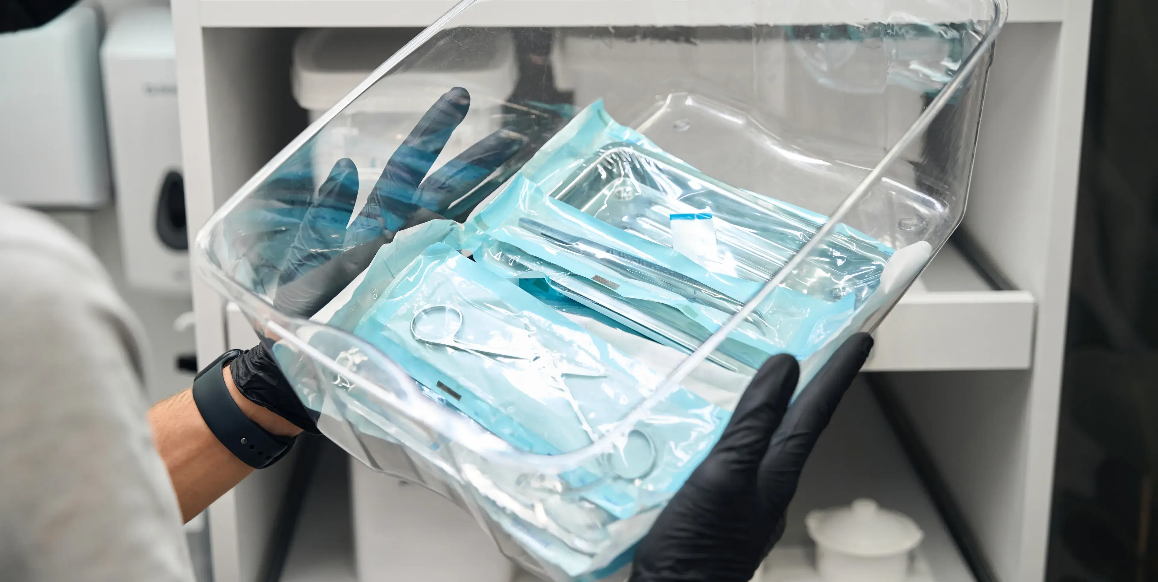 Person in black gloves holds a clear bin containing sterilized dental tools in blue packages.