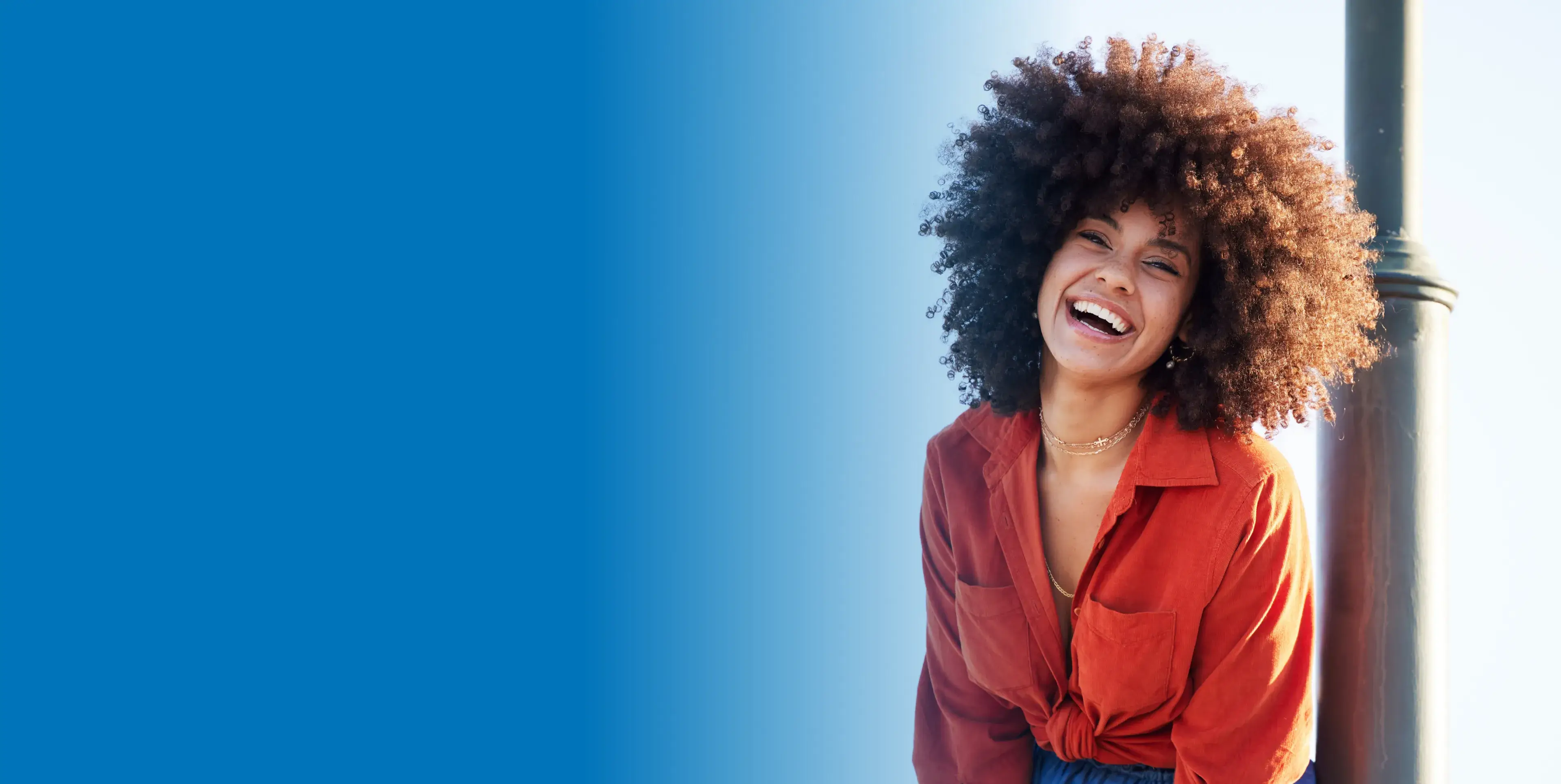 A person with curly hair is smiling broadly, wearing an orange shirt in bright sunlight.