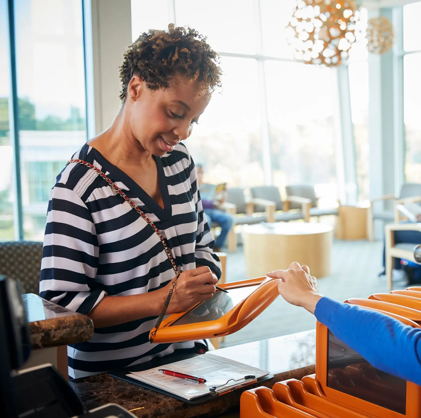 A woman signs papers at a hotel reception desk while holding a bright orange folder.