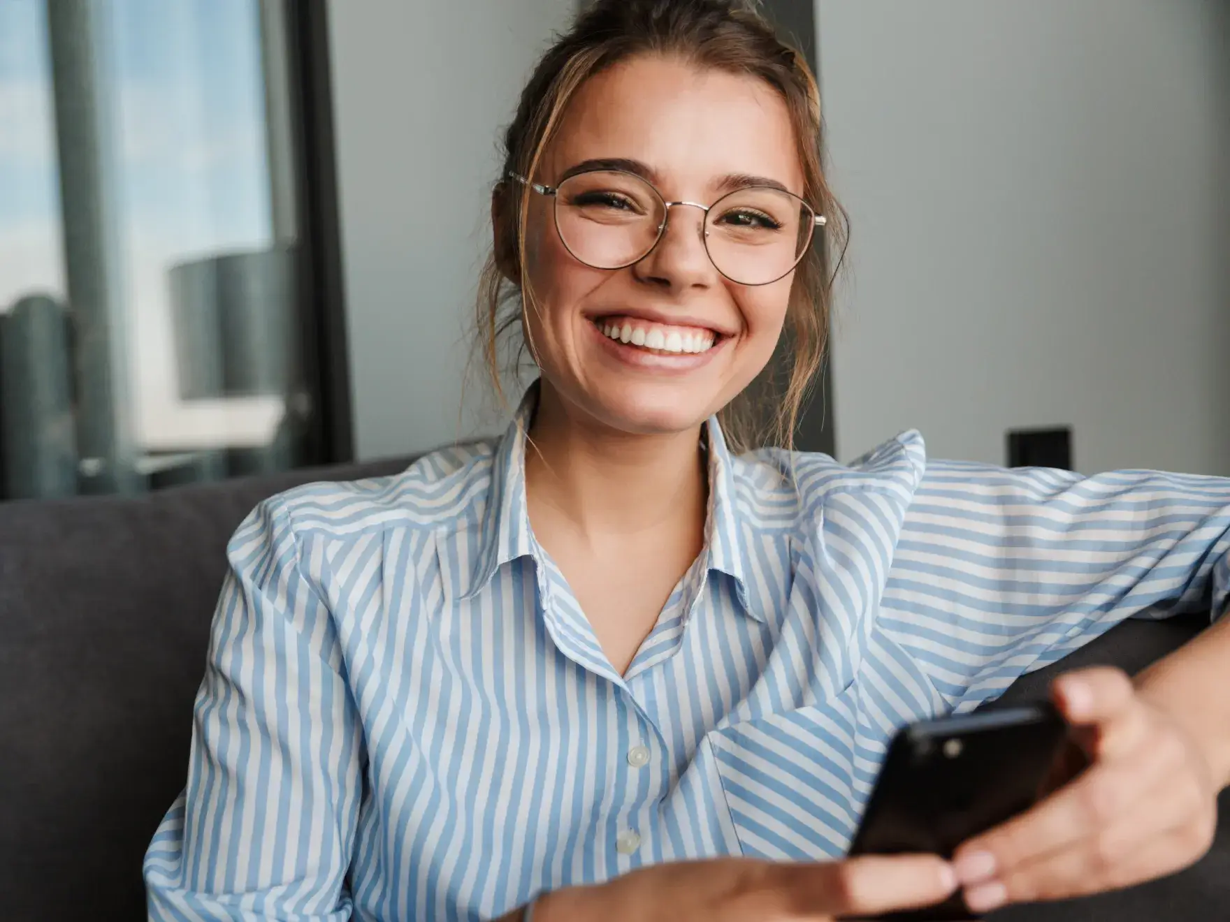 A woman in a striped shirt sits on a couch, smiling and holding a smartphone.