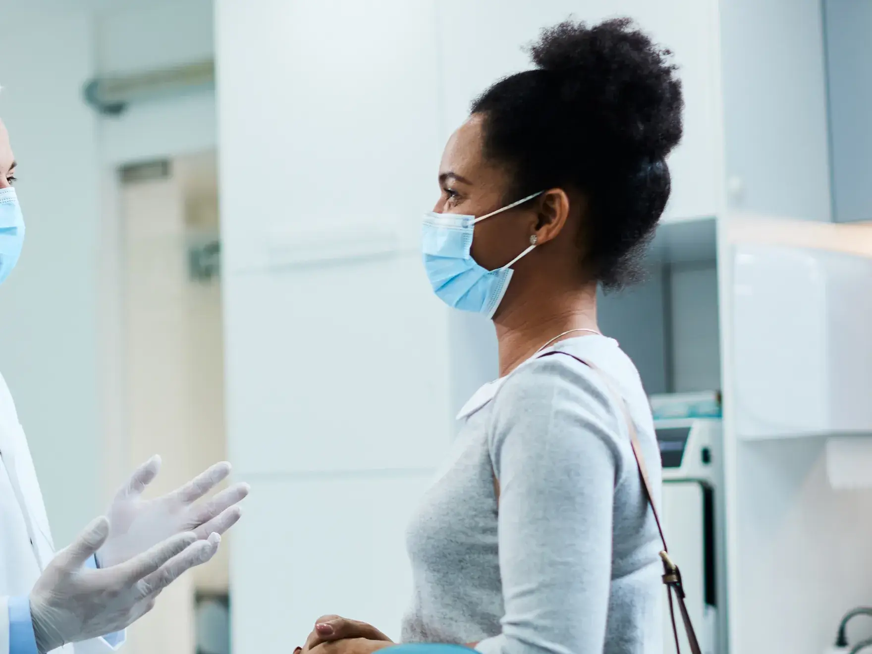 A doctor wearing gloves talks with a woman wearing a mask in a medical office.