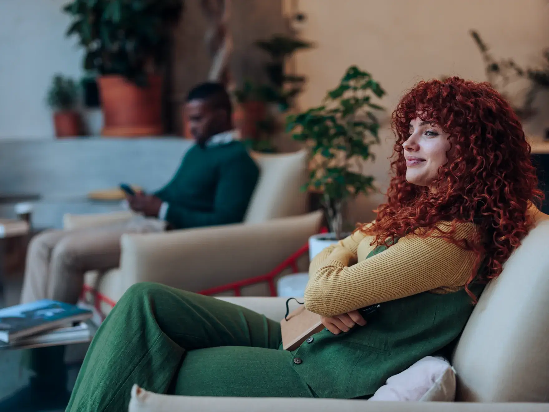 Woman with curly red hair sits on a couch, smiling, while a man uses a phone nearby.