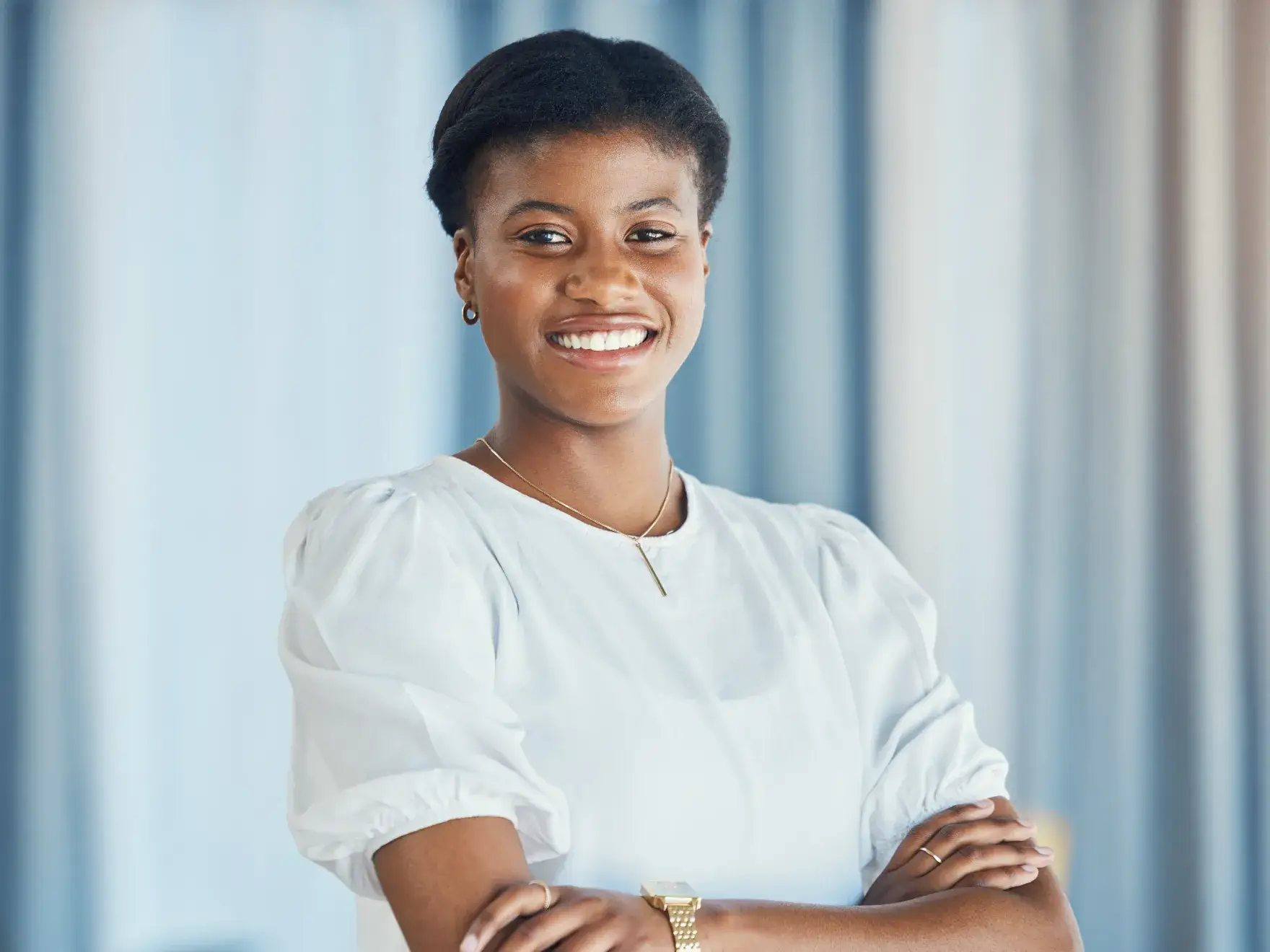 A person smiling confidently with arms crossed, wearing a white blouse against a blurred background.