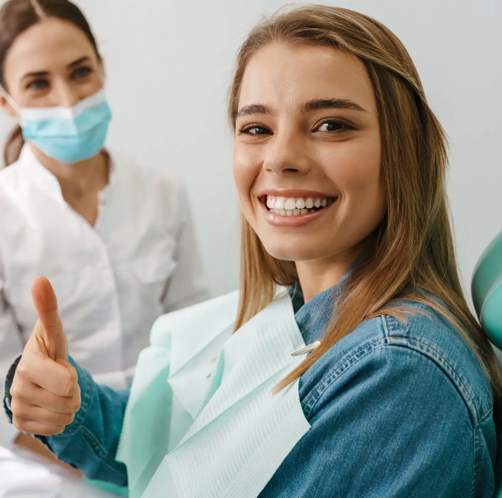 A woman sitting in a dentist's chair gives a thumbs-up, with a dental professional nearby.