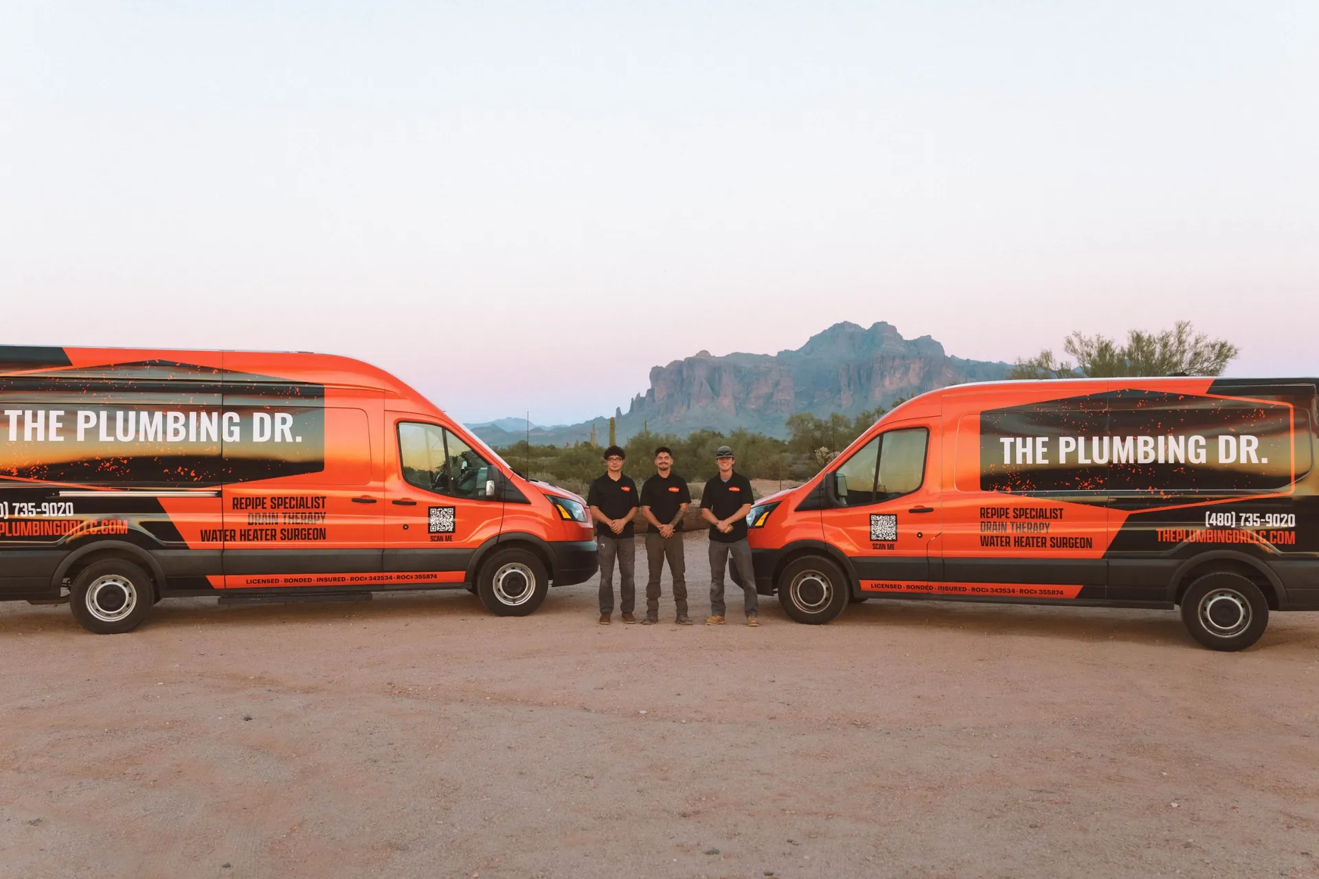 Plumbing Dr. technicians standing between two service vans at sunset near the Superstition Mountains