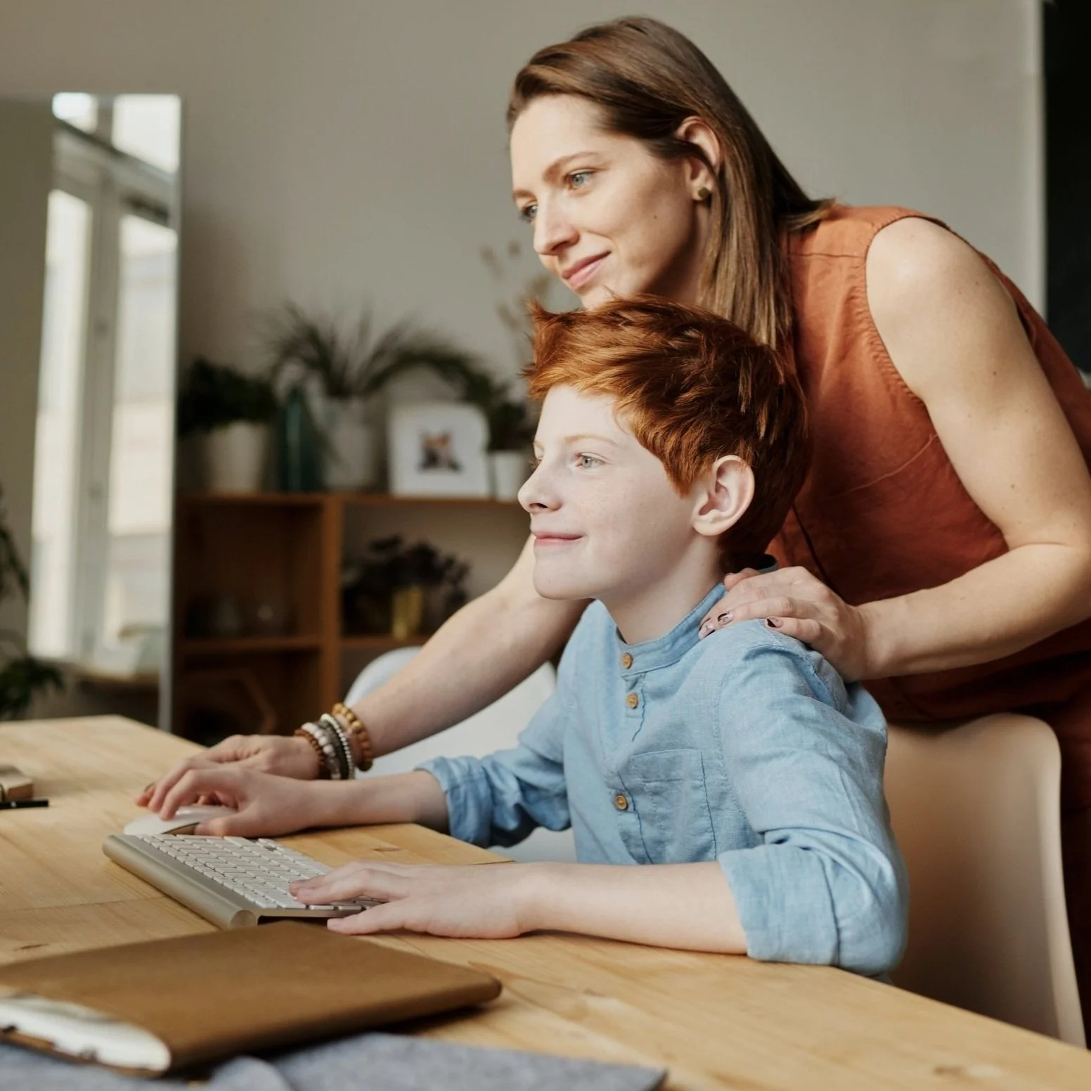 A woman with brown hair in a sleeveless orange top stands behind a red-haired boy in a blue shirt who is using a computer keyboard, both looking at the screen with focused expressions in a cozy room with plants and shelves in the background.