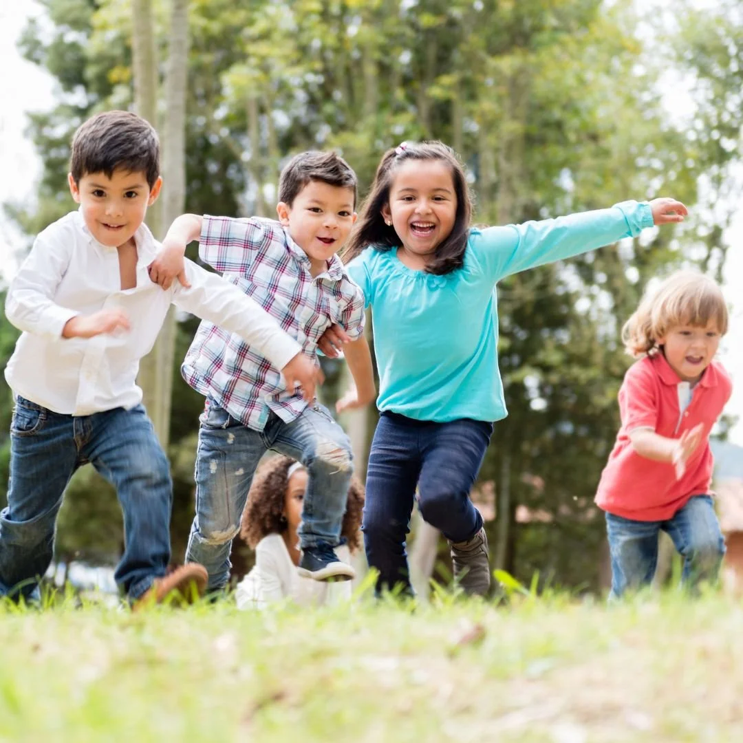 Four children running outdoors on grass with trees in the background, smiling and appearing joyful during playtime.