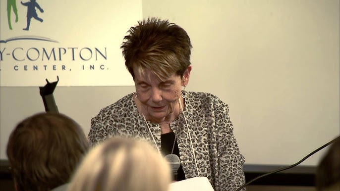 An older woman with short hair and leopard-print jacket speaking into a microphone at an event, with a sign partially visible behind her.