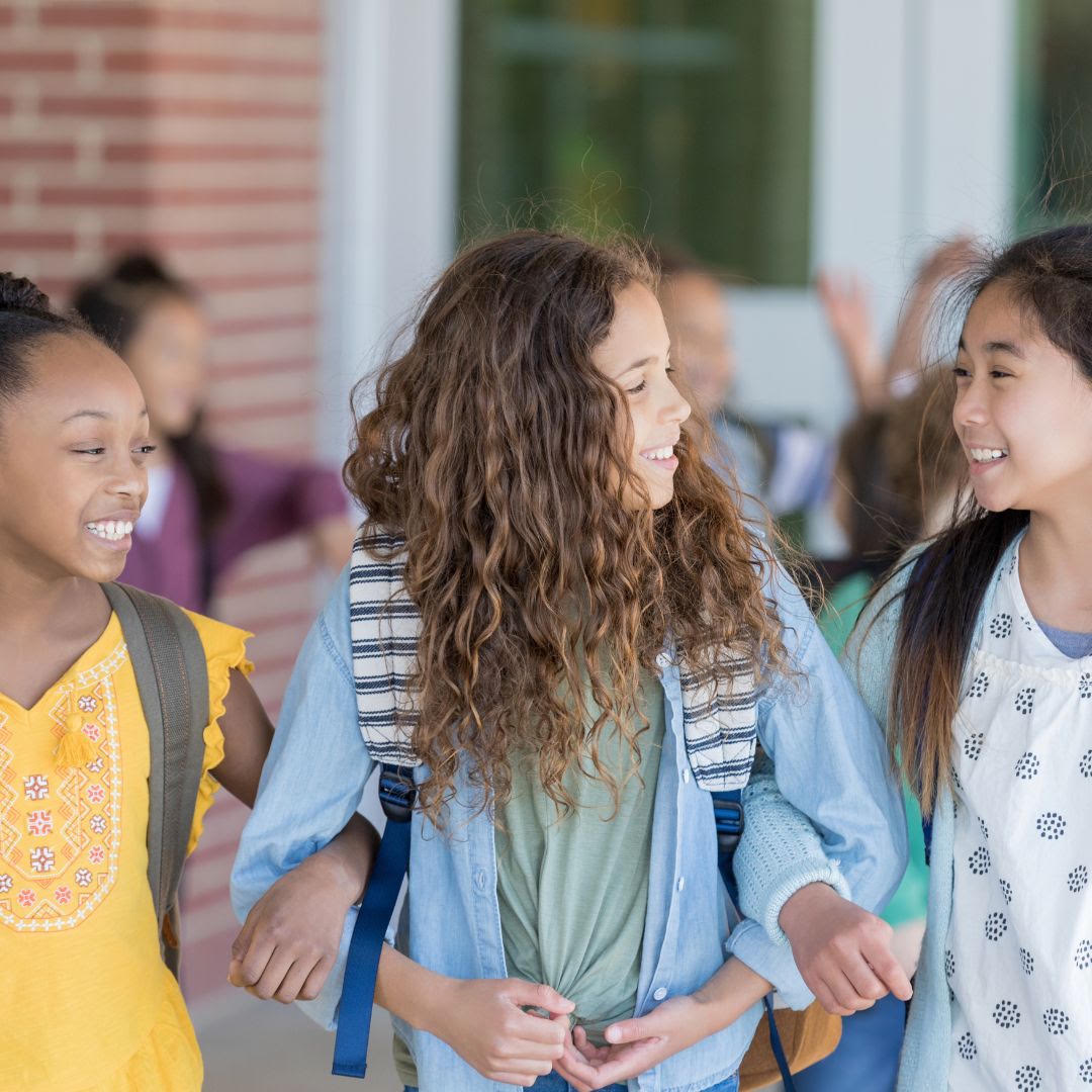 Three smiling girls walking outdoors, linked arm in arm, carrying backpacks, with a school building in the background.