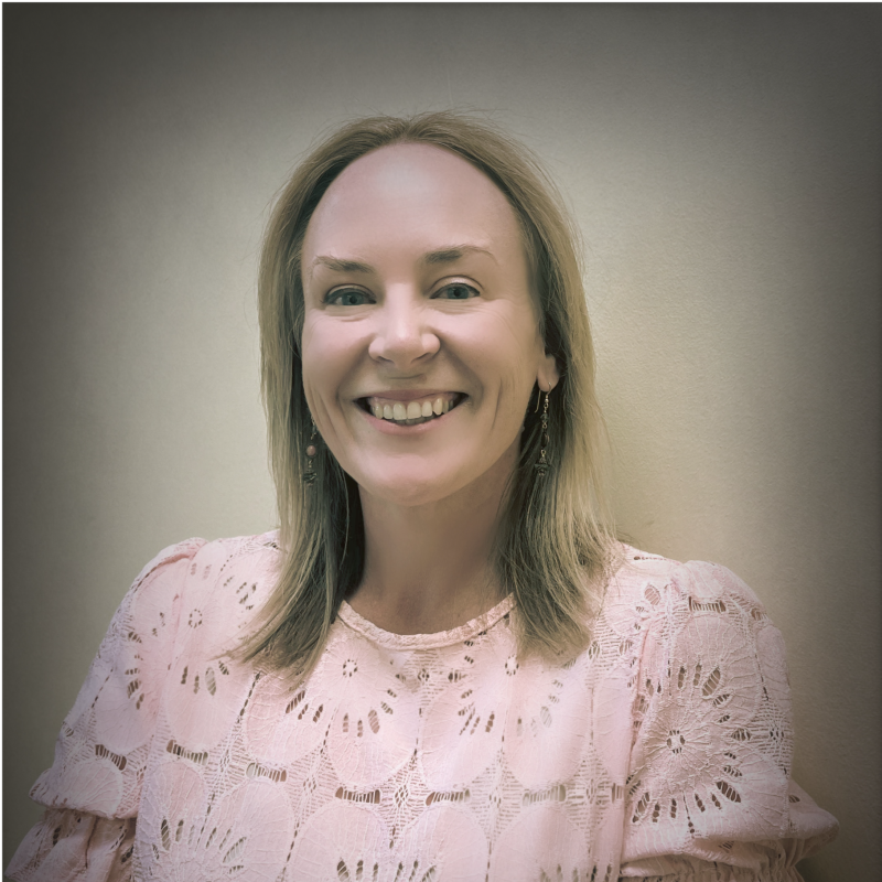 Smiling woman with shoulder-length blonde hair wearing a pale pink lace blouse and dangling earrings against a neutral background.