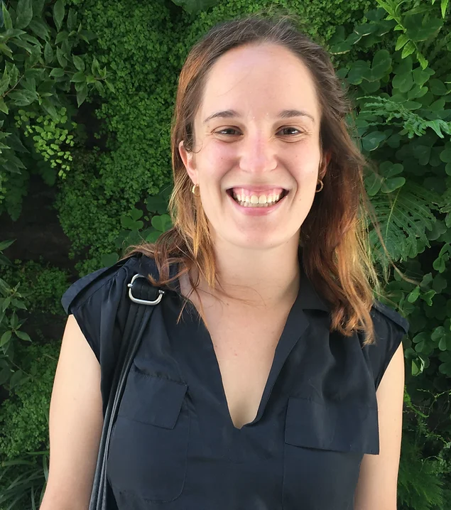 Smiling woman with light skin and brown hair wearing a navy sleeveless top standing in front of green leafy plants.