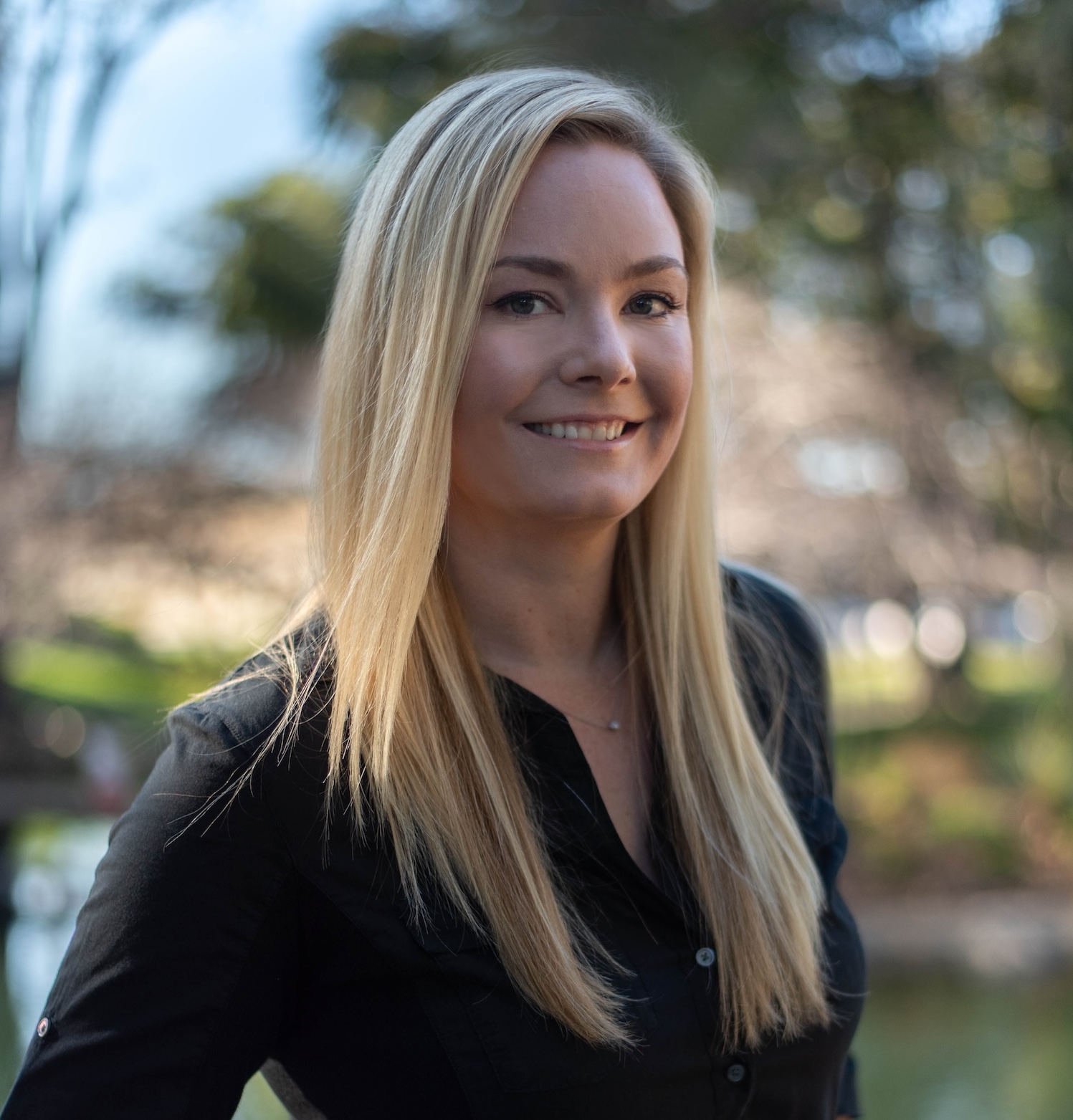 Portrait of a smiling woman with long straight blonde hair wearing a black button-up shirt, with a blurred outdoor background of trees and sunlight.
