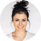 Smiling woman with dark hair tied up in a messy bun wearing a white top against a light background.