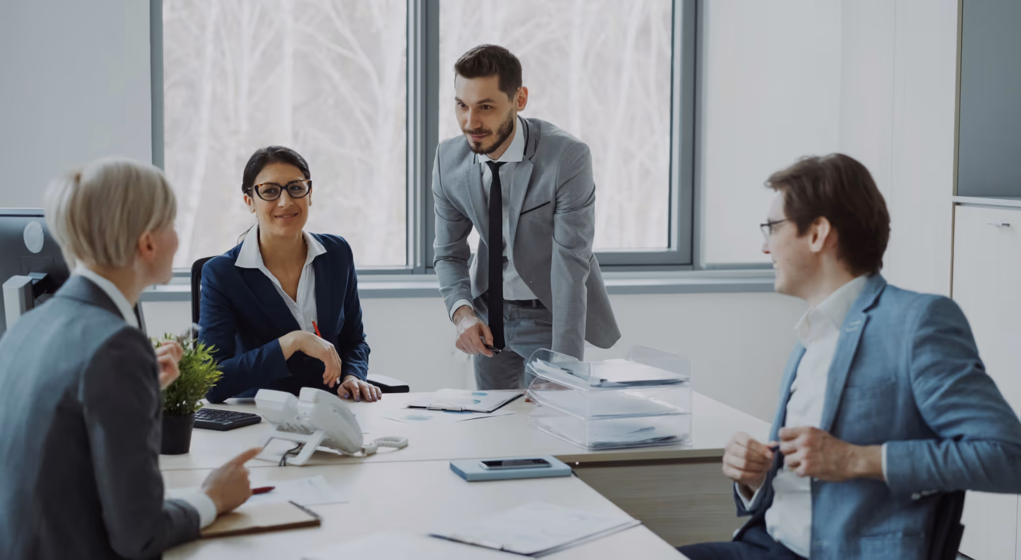 Four business professionals engaged in a meeting around a desk with documents and office supplies.